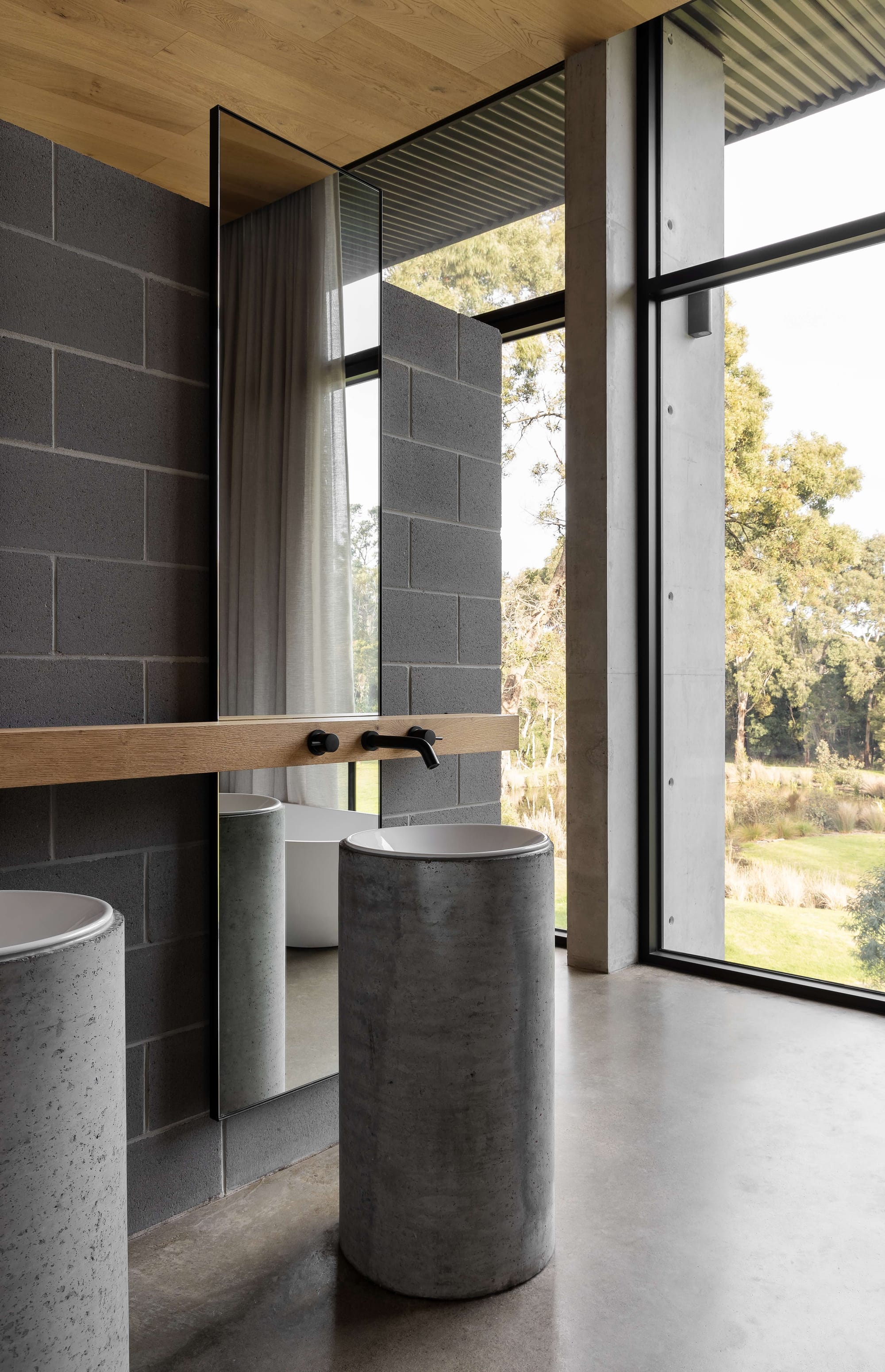 Merricks House by Aktis Architects. Photography by Timothy Kaye. Modern bathroom with cylindrical concrete basins, full-height mirror, blockwork walls, and expansive windows overlooking a natural landscape.