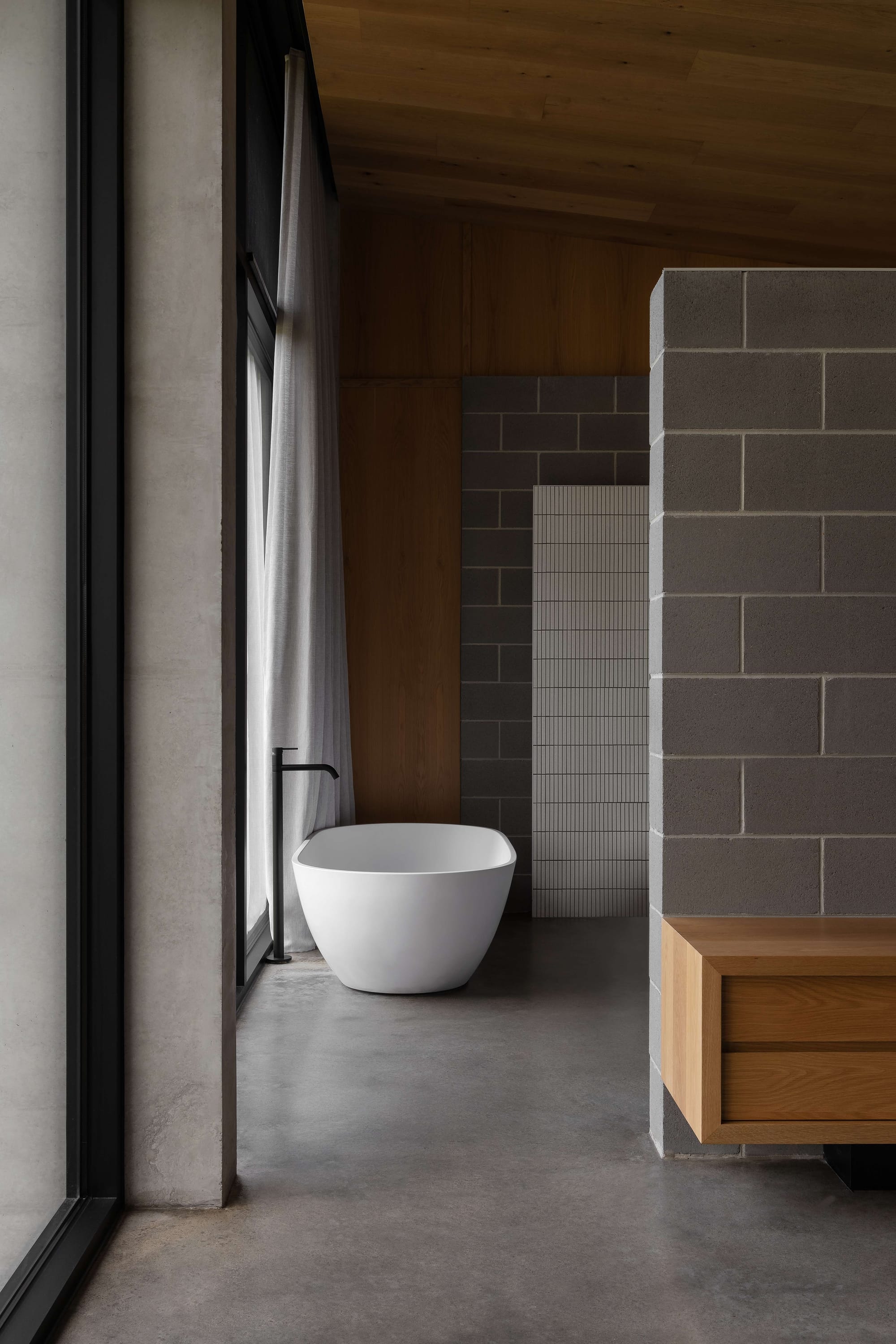 Merricks House by Aktis Architects. Photography by Timothy Kaye. Serene bathroom with freestanding white tub, grey concrete block walls, minimalist black tapware, and full-height windows with sheer curtains.