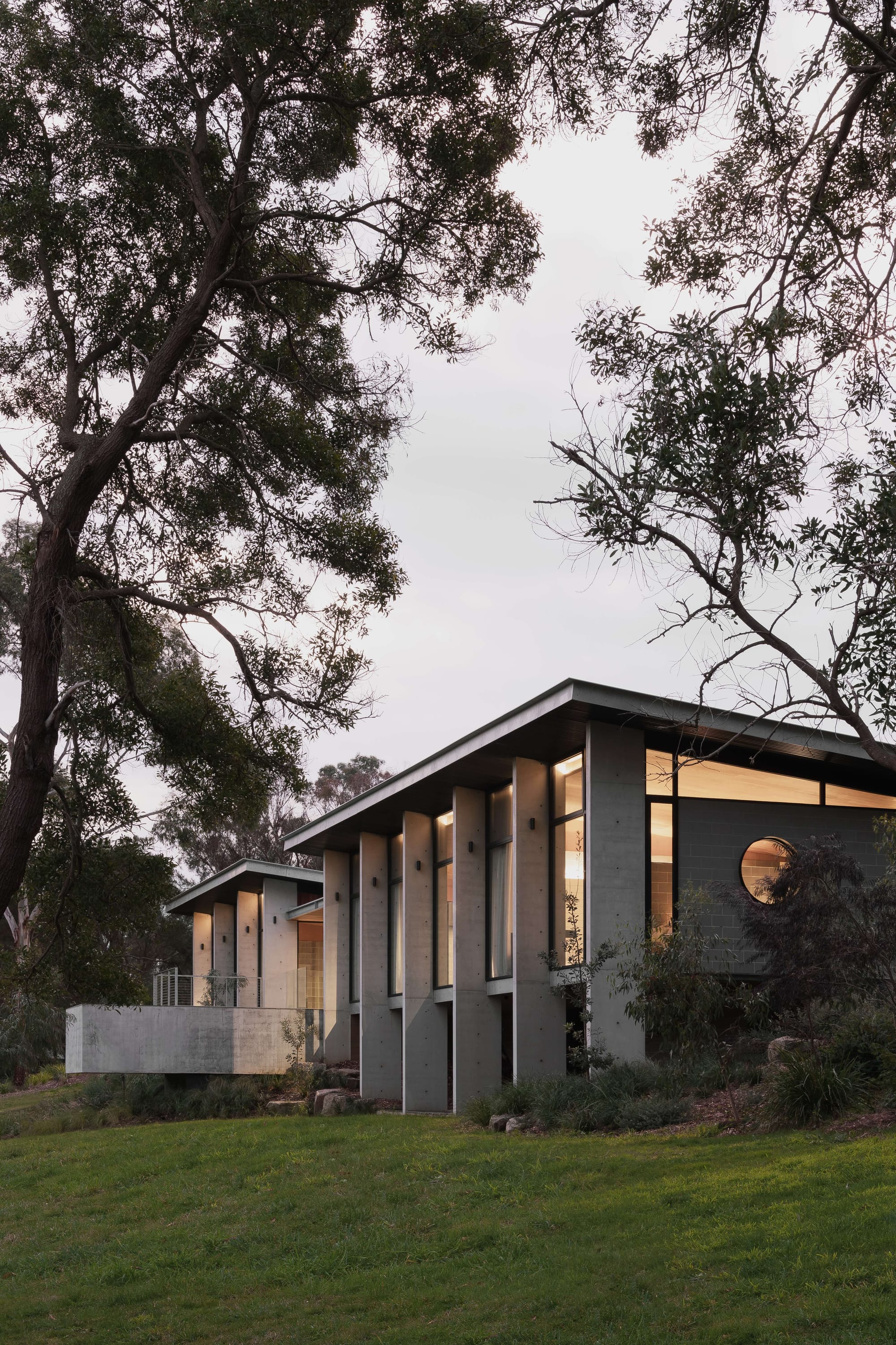 Merricks House by Aktis Architects. Photography by Timothy Kaye.Modern concrete house with narrow vertical fins and clerestory glazing, framed by native trees and evening light.