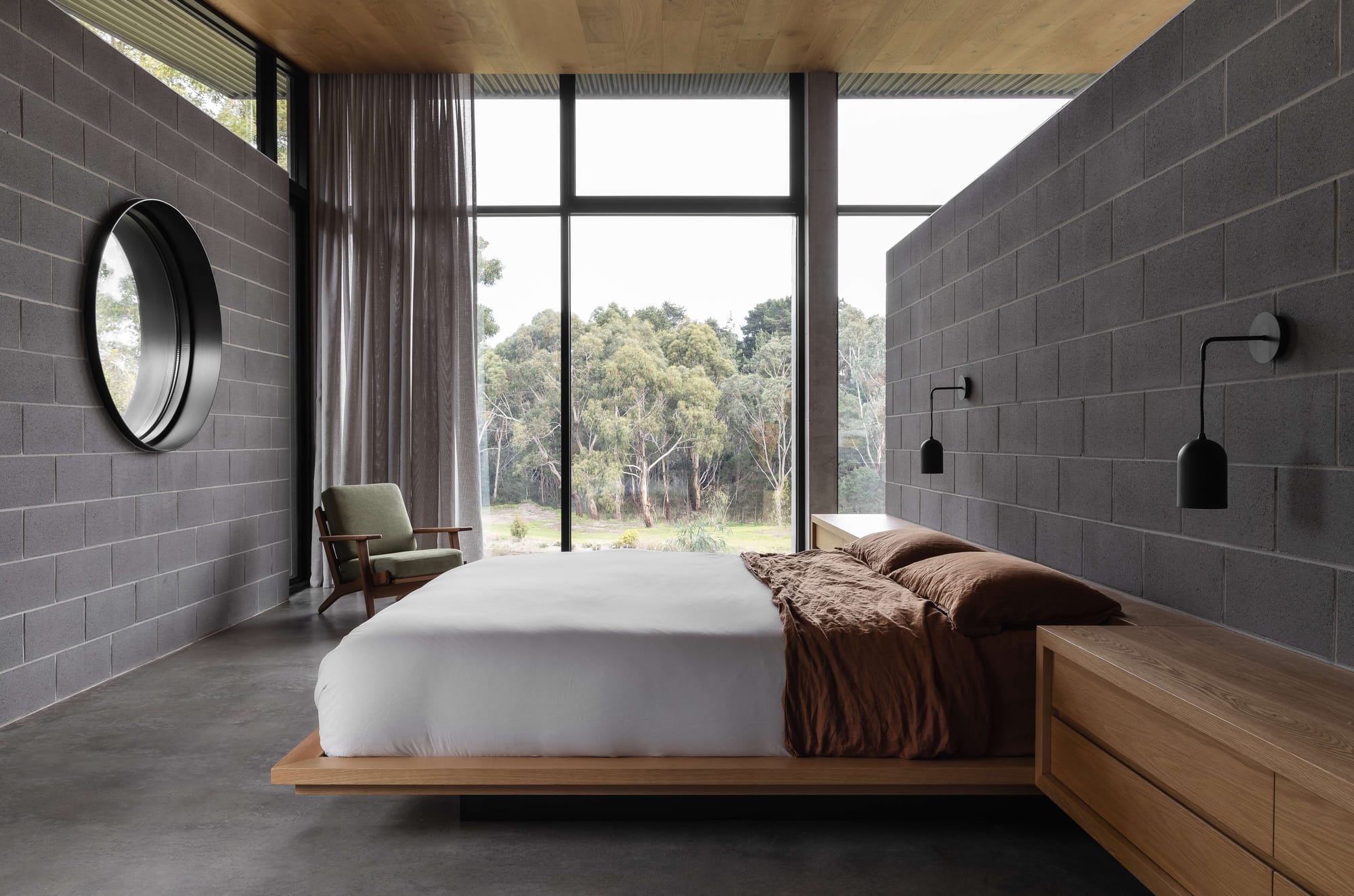 Merricks House by Aktis Architects. Photography by Timothy Kaye. Minimalist bedroom with concrete block walls, timber floating platform bed, large picture window framing forest views, and a circular wall mirror.