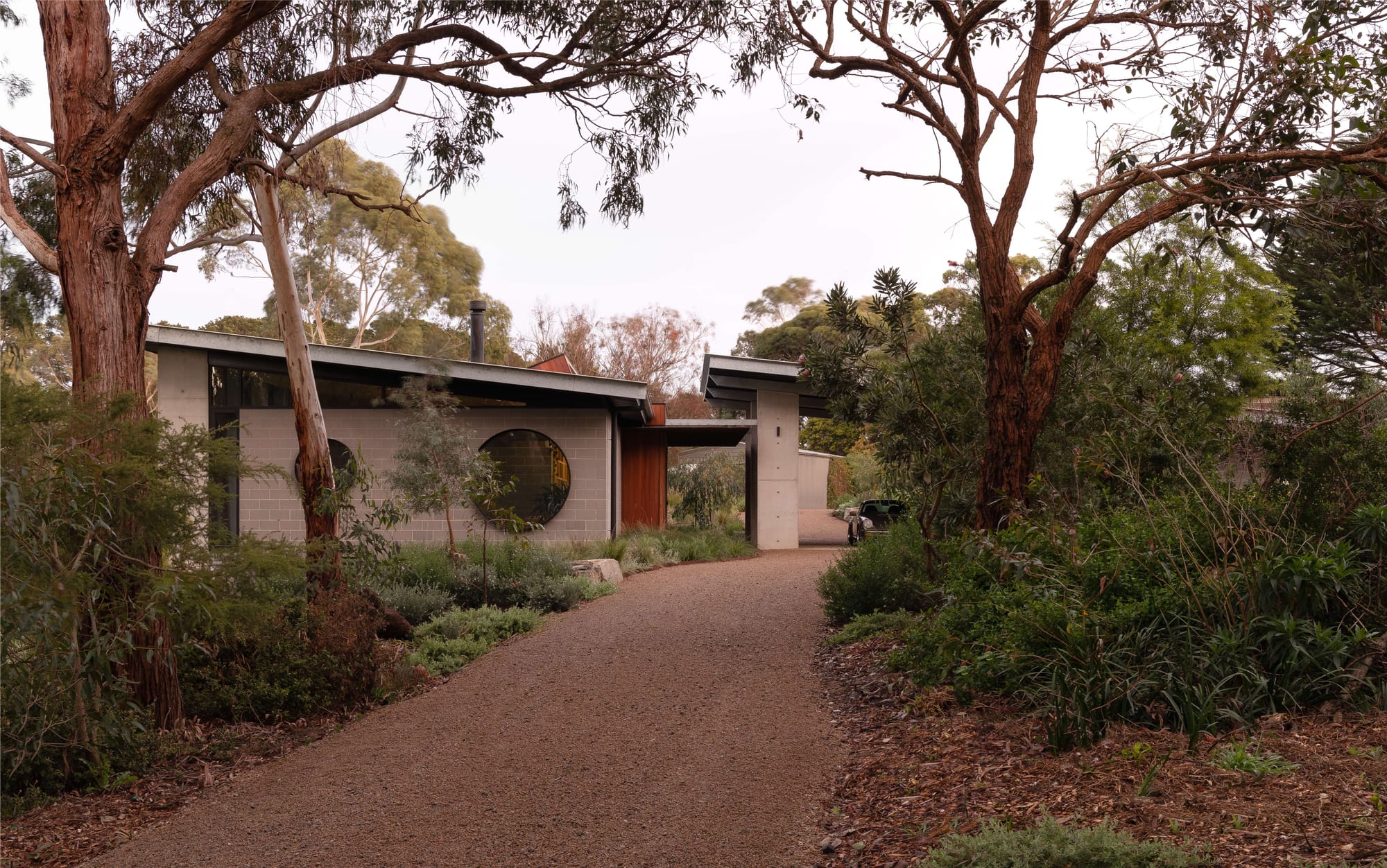 Merricks House by Aktis Architects. Photography by Timothy Kaye. Exterior view of a modern house nestled in native Australian bushland, featuring grey blockwork walls, a distinctive round window, and a low-sloped metal roof surrounded by tall eucalyptus trees.