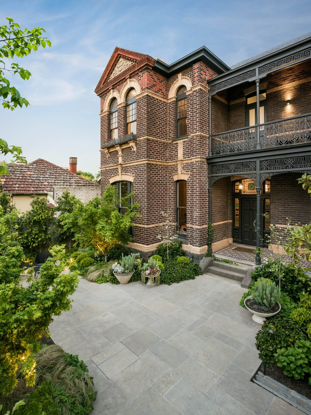 Armadale by Myles Baldwin Design. Photographer undisclosed. Dark double storey Victorian terrace home with paved driveway and lush garden beds.