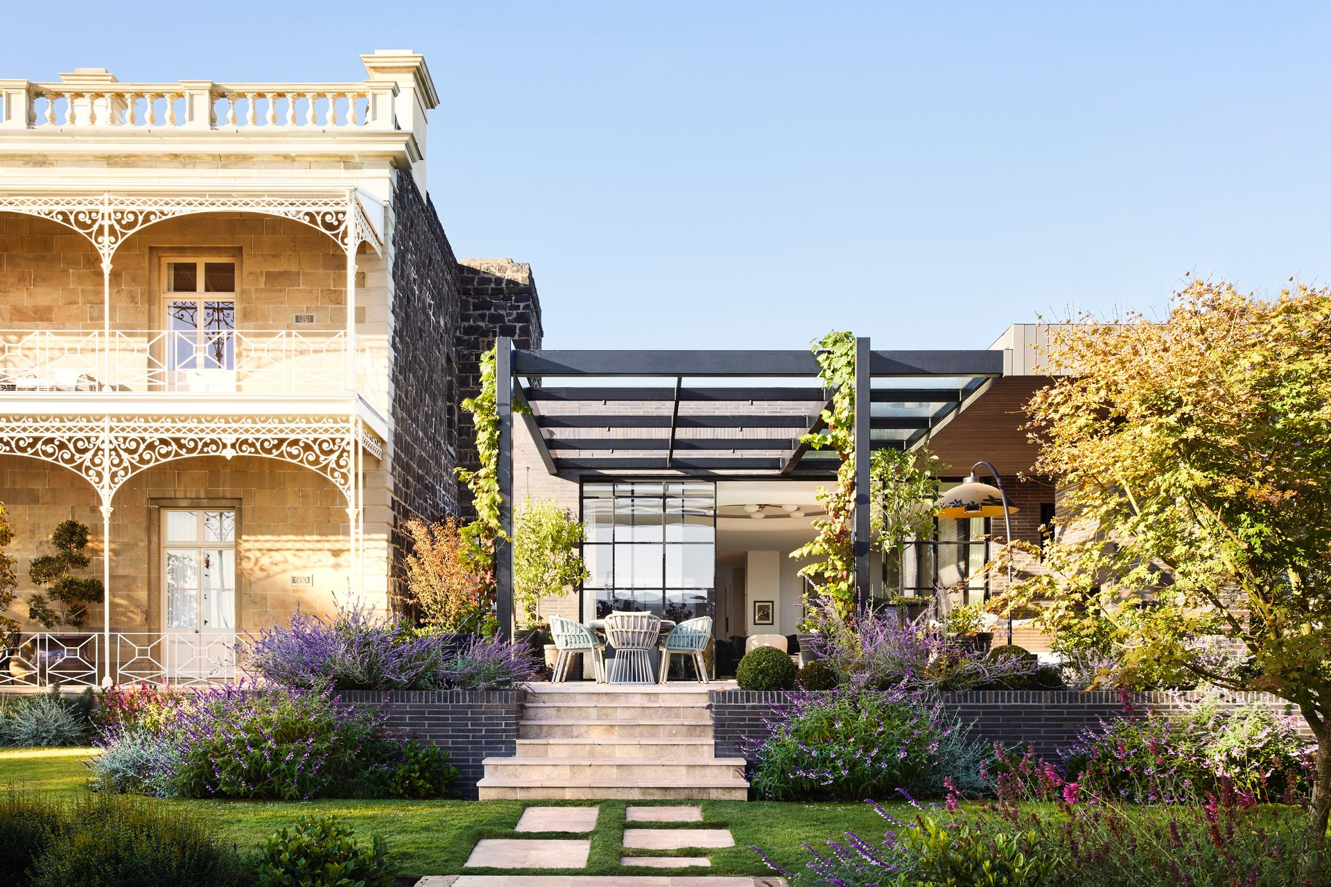 Geelong, Victoria by Myles Baldwin Design. Photographer undisclosed. Patio alongside double storey Victorian home with colourful gardens and grass.