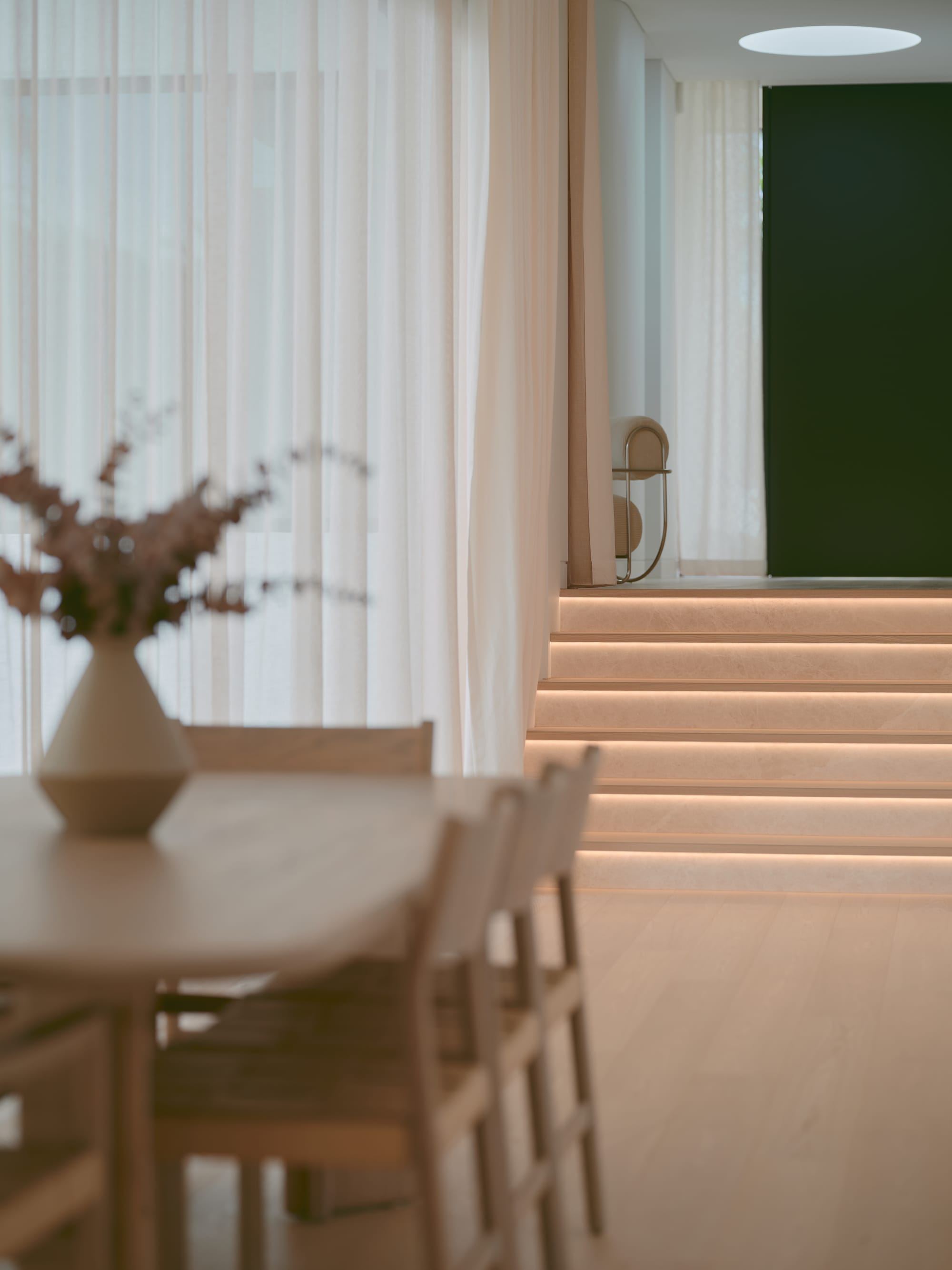 Sole House by 868 Architects. Photography by Martin Siegner. Softly lit stairway with concealed lighting leads from a serene dining area framed by sheer curtains to a hallway featuring a deep green accent wall.