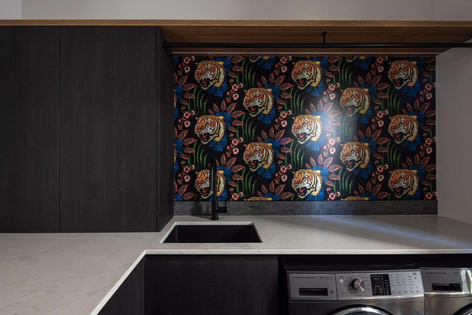 Queenscliff 08 by Watershed Architects. Photography by Simon Whitbread. Laundry with black cupabords, white countertops and wallpaper splashback, featuring a tigers head in front of purple and blue flowers and green leaves.