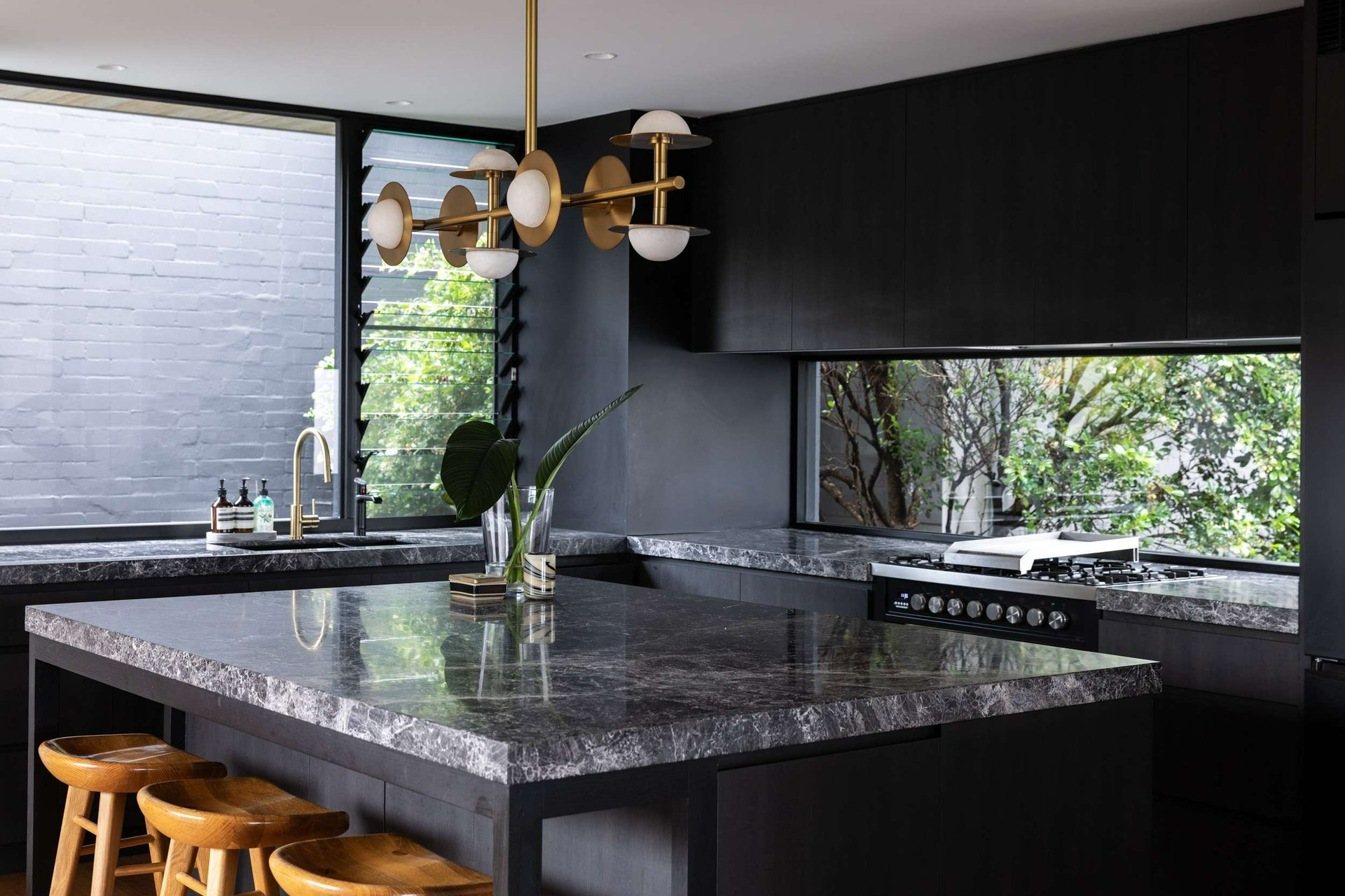 Queenscliff 08 by Watershed Architects. Photography by Simon Whitbread. Dark kitchen with black cabinetry, dark stone countertop, gold pendant light and glass window splash back.