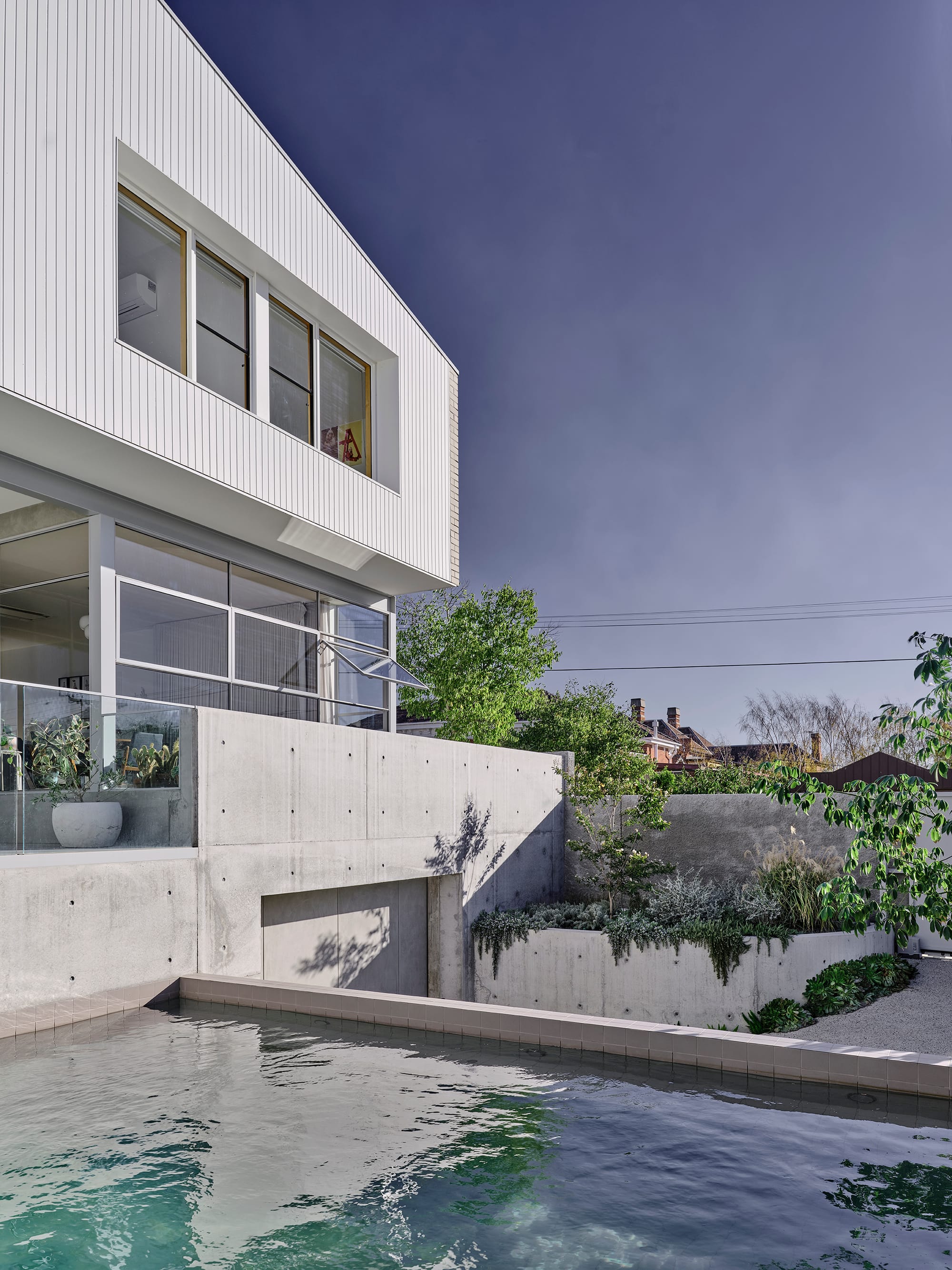 Salmon House by True Story. Photography by Dean Bradley. White gable-roofed home with a concrete base and glass balustrade, overlooking a turquoise pool and outdoor dining area.