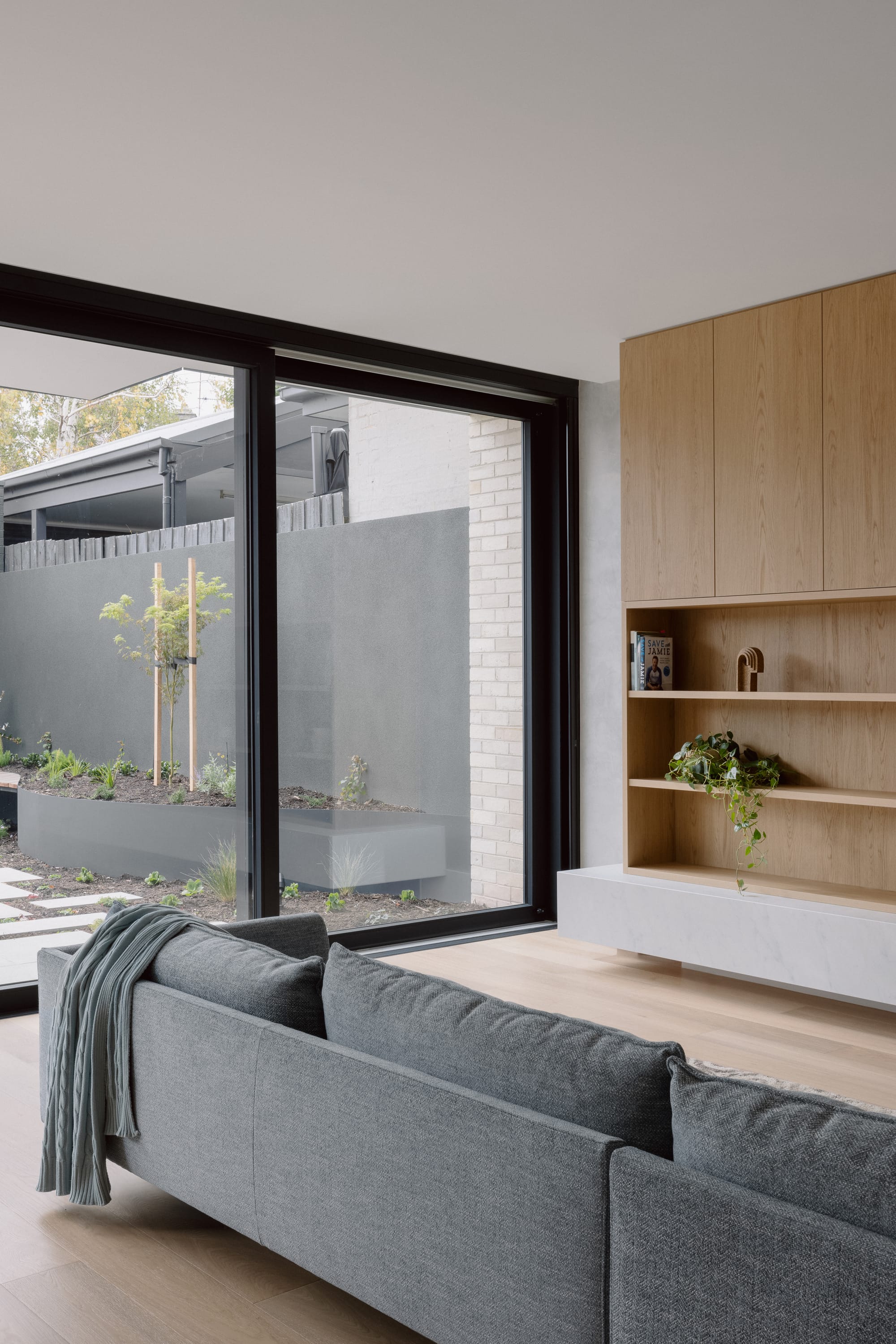 Auburn Residence by Chan Architecture. Photography by Elise Scott. Living space with timber cabinetry, flooring and grey couch opening onto external courtyard through black framed glass sliding doors.
