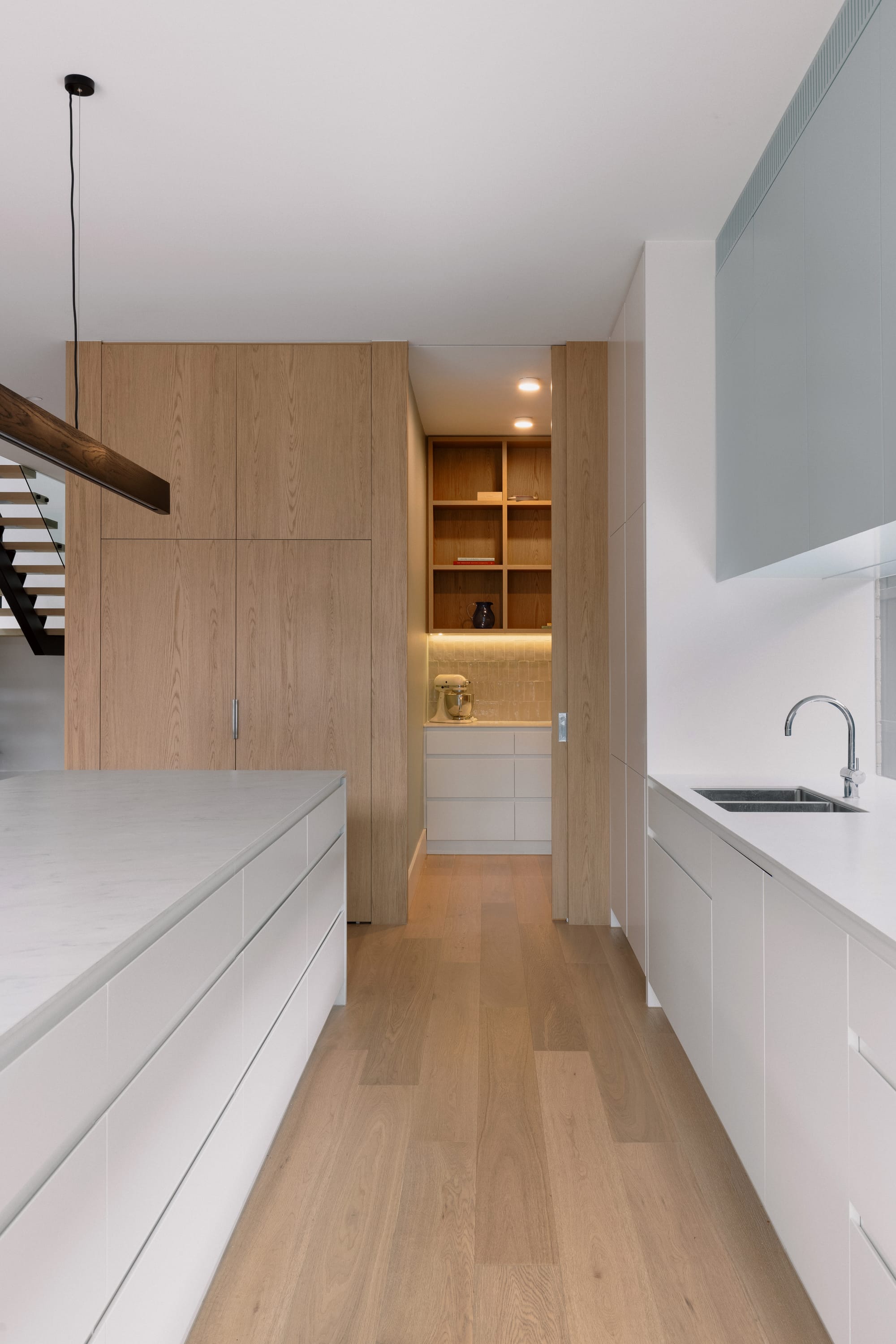 Auburn Residence by Chan Architecture. Photography by Elise Scott. Open plan kitchen with white cabinets and benchtops, timber flooring and pantry and scullery with overhead cupboards and tiled splashback.