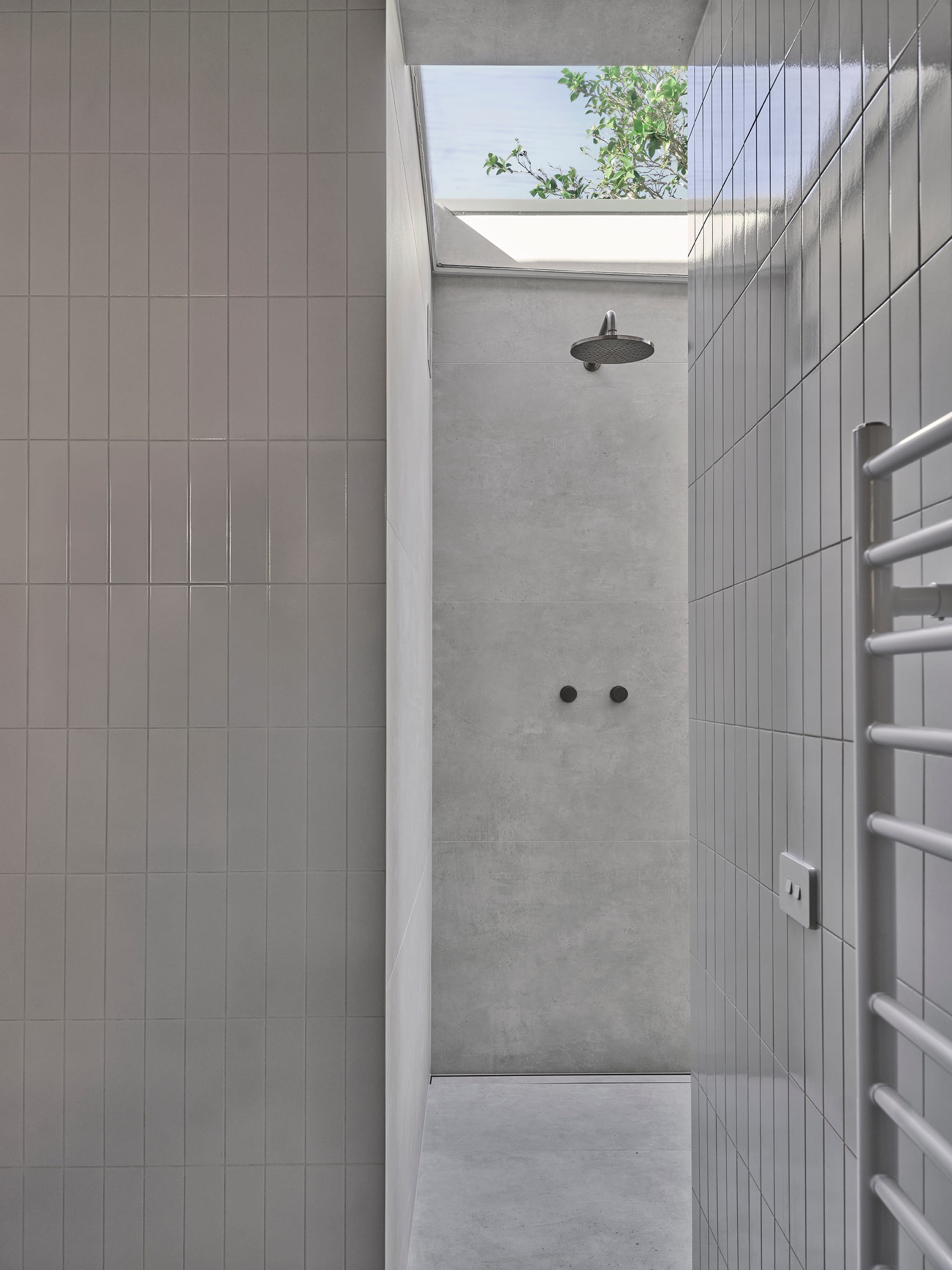 Salmon House by True Story. Photography by Dean Bradley.Minimalist shower nook with concrete and vertical tiles, lit from above by a skylight framing tree branches.