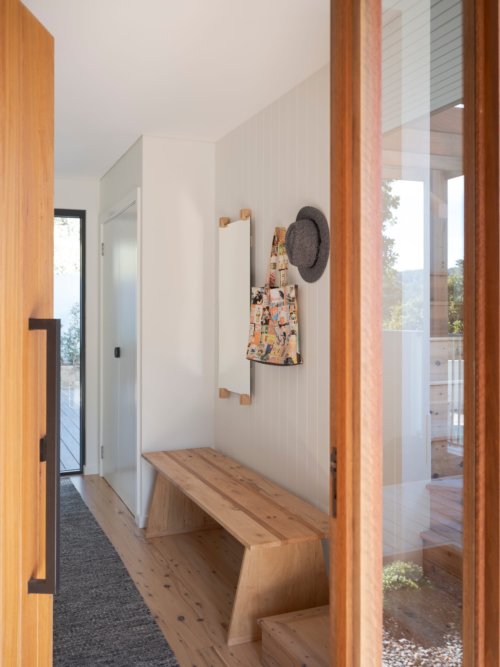 Three Points House by BIJL Architecture. Photography by Tom Ferguson.View through an open timber door into a light-filled entryway with a timber bench, wall hooks, a hat, and a patterned tote bag hanging against vertical white paneling.