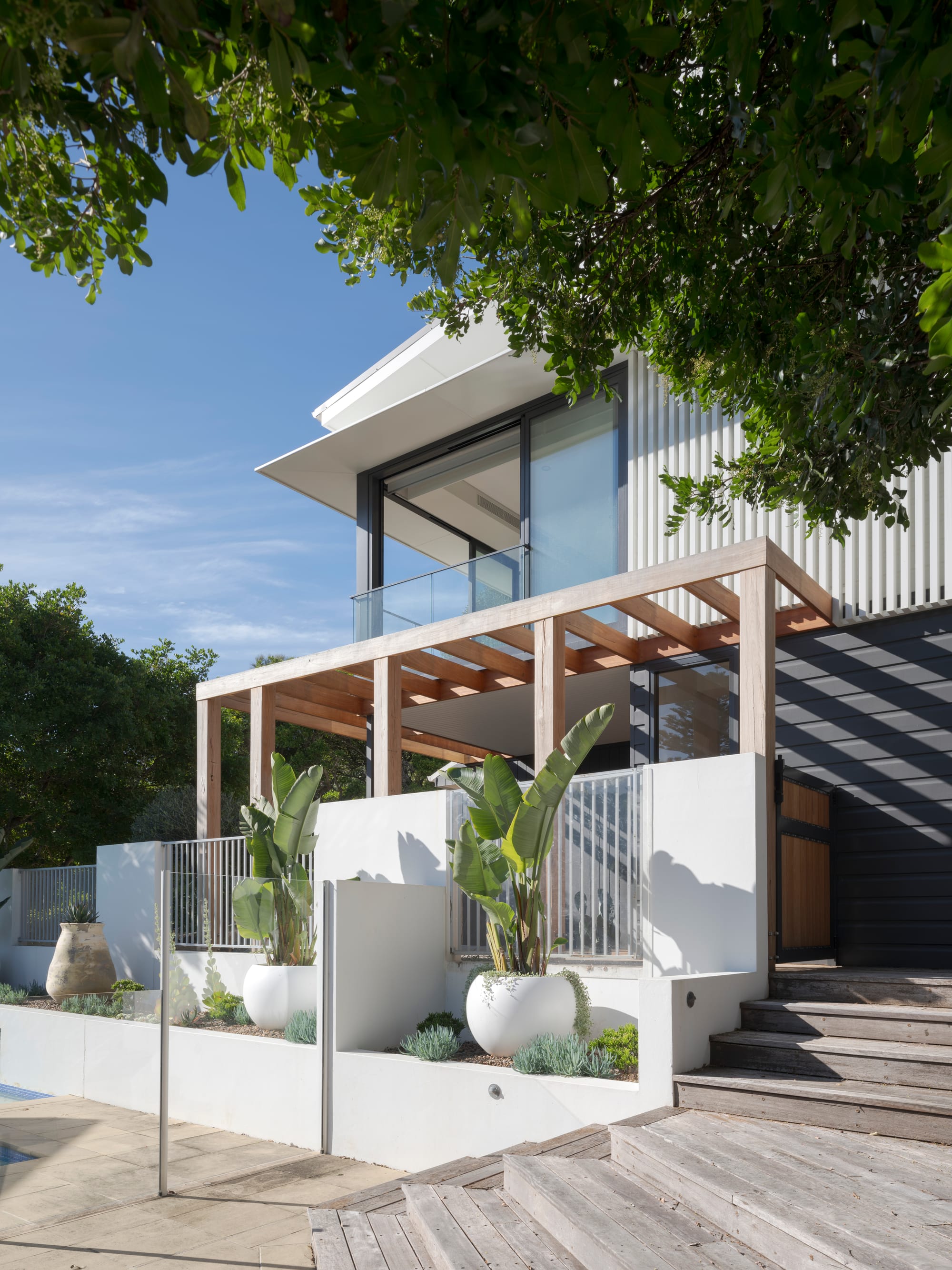 Three Points House by BIJL Architecture. Photography by Tom Ferguson.Contemporary home with a glass balcony, timber pergola, and tiered landscaping featuring white planter pots, lush greenery, and a timber deck beside a pool area.