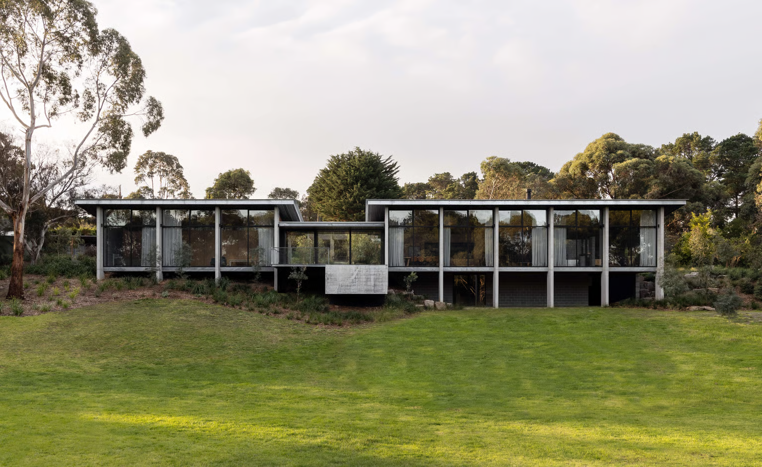 Merricks House by Aktis Architetcs. Photographed by Timothy Kaye. Modern pavilion-style home set on a sloping site, featuring a linear form with full-height glazing, concrete columns, and a central entry bridge framed by native landscaping.