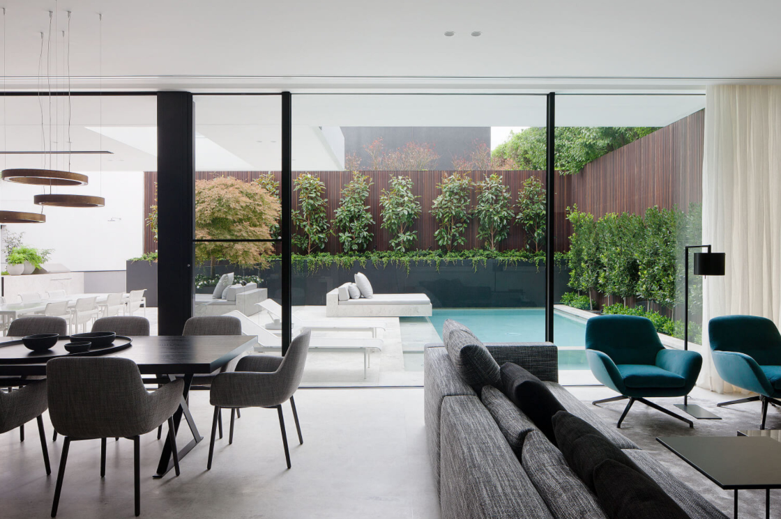 Kent House Toorak by Emma Tulloch Architects. Photography by Shannon McGrath. Sophisticated open-plan living and dining area with floor-to-ceiling glass doors opening to a modern courtyard featuring a pool, vertical garden, and minimalist outdoor seating.