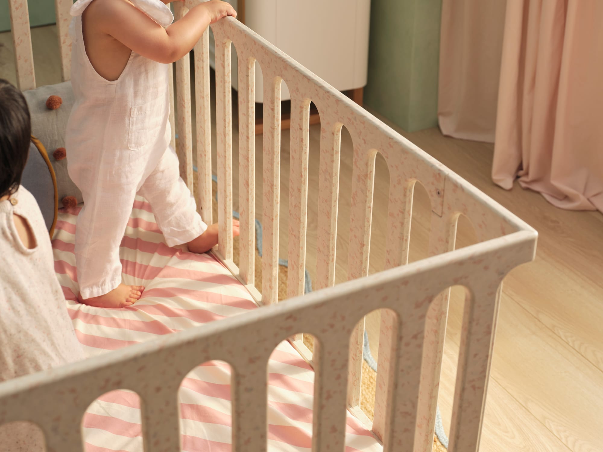 Archie Cot by Seena x Defy in Dalmation Pink. White and pink cot with pink and white striped mattress cover in room with timber floors, green walls and pink curtains.