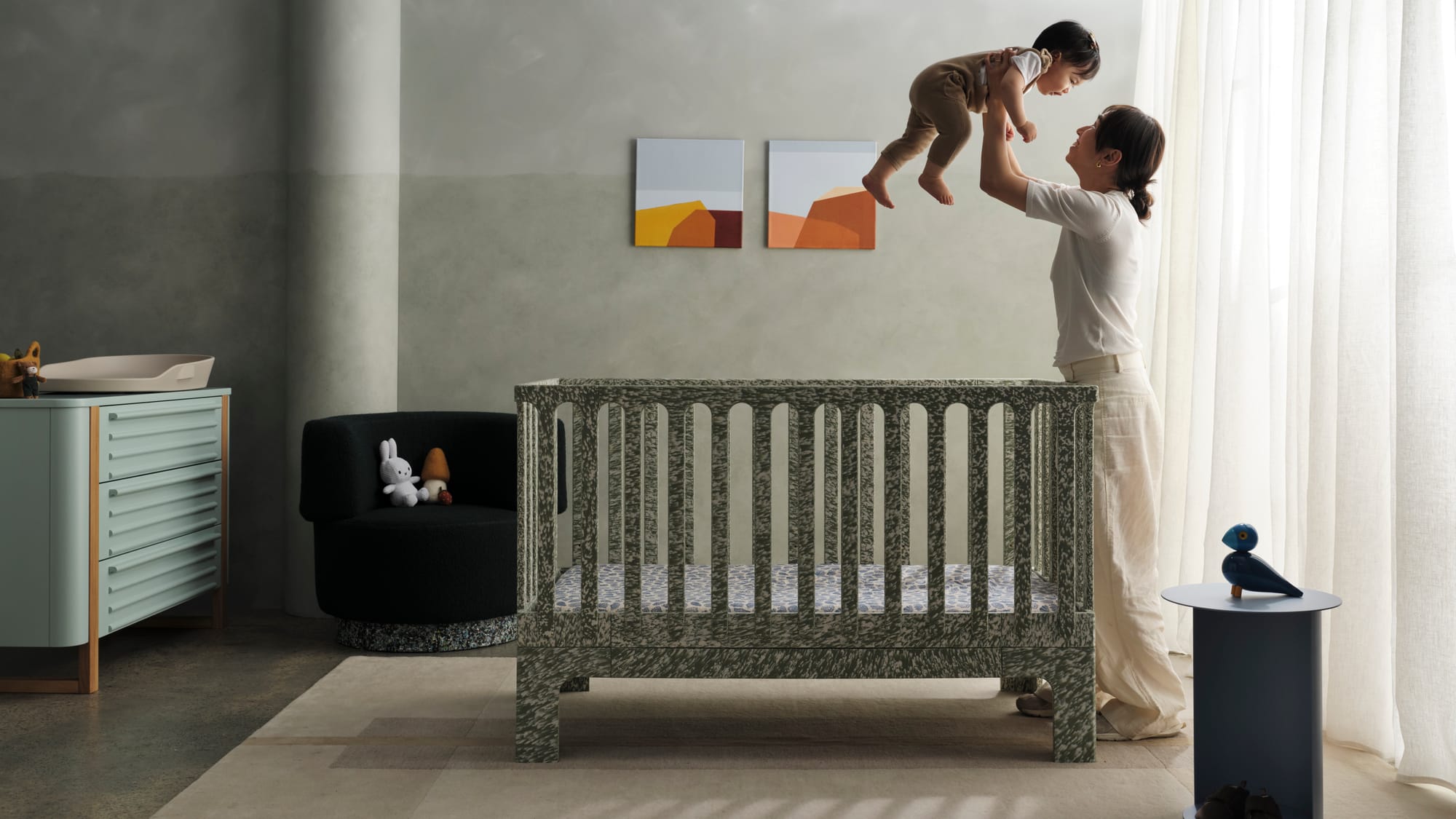 Archie Cot by Seena x Defy in Sage Spray. Woman holding toddler above her bed, above green and white speckled cot in room with concrete walls, floors and white rug.
