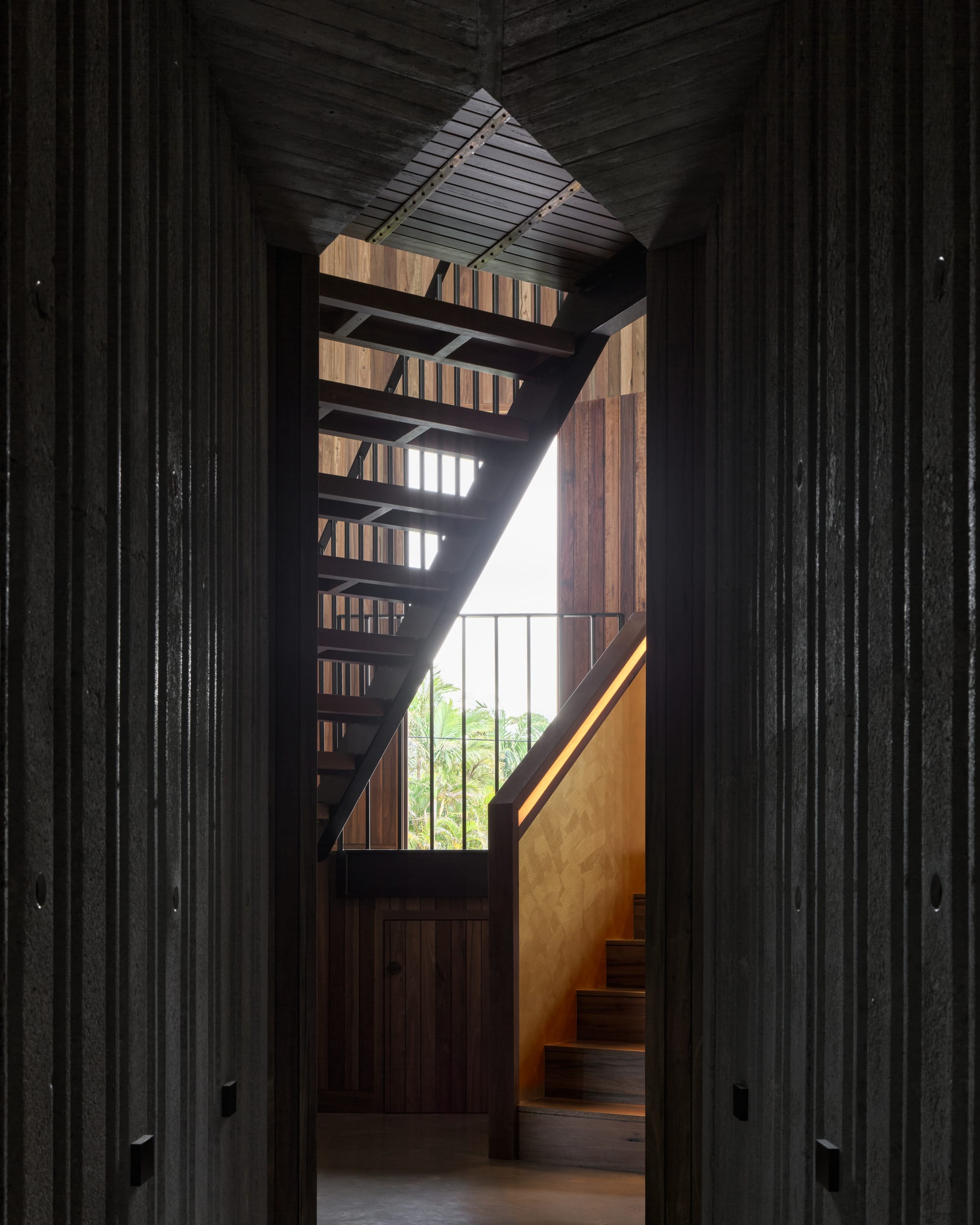 Cake House by Alexander Symes Architect. Photography by Barton Taylor. Black timber clad walls on left and right-side framing timber staircase in front of a window, looking out over tropical plants.