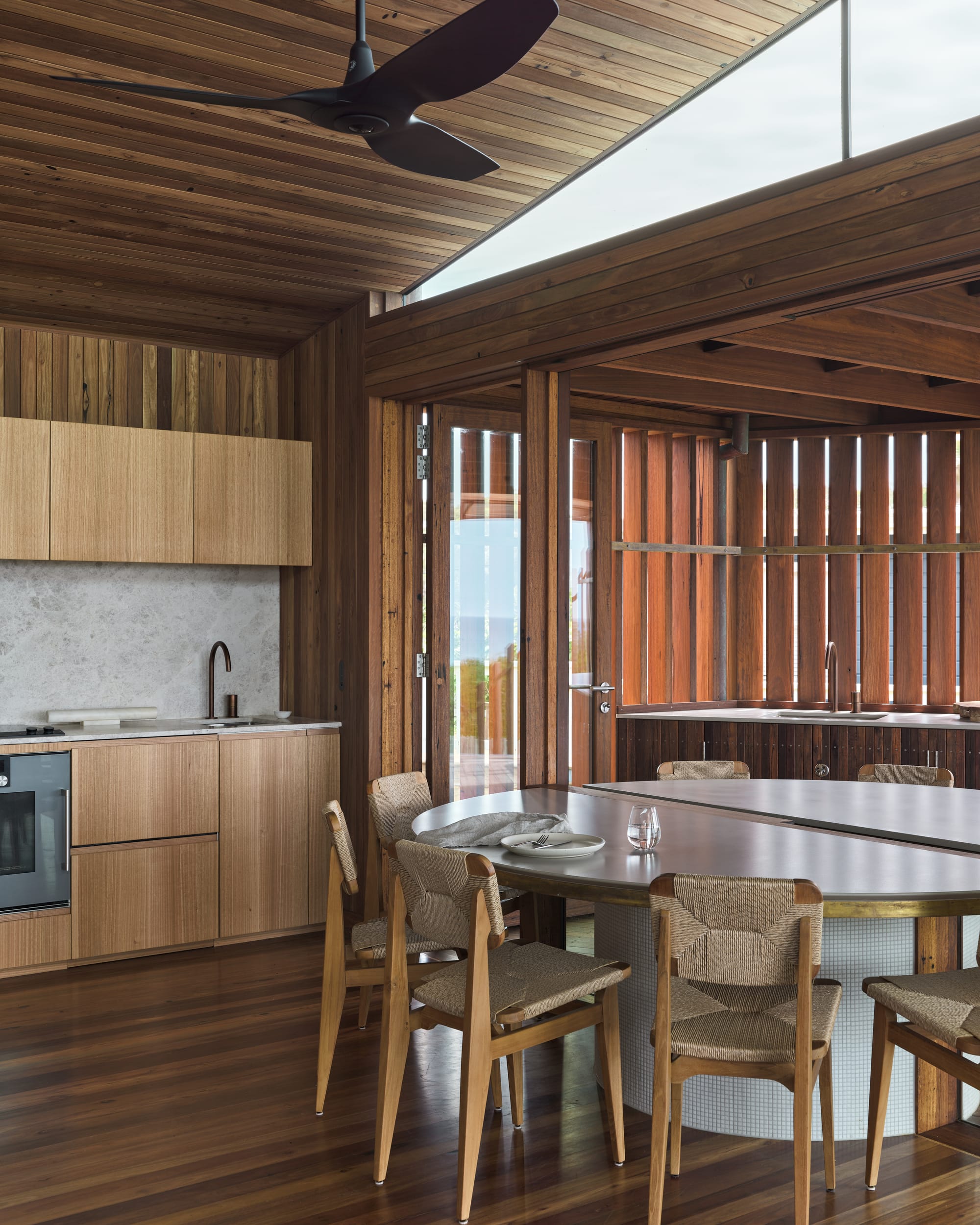 Cake House by Alexander Symes Architect. Photography by Barton Taylor. Kitchen and dining space with timber walls, floors, ceiling and cabinetry, and a white round dining table with rattan chairs.