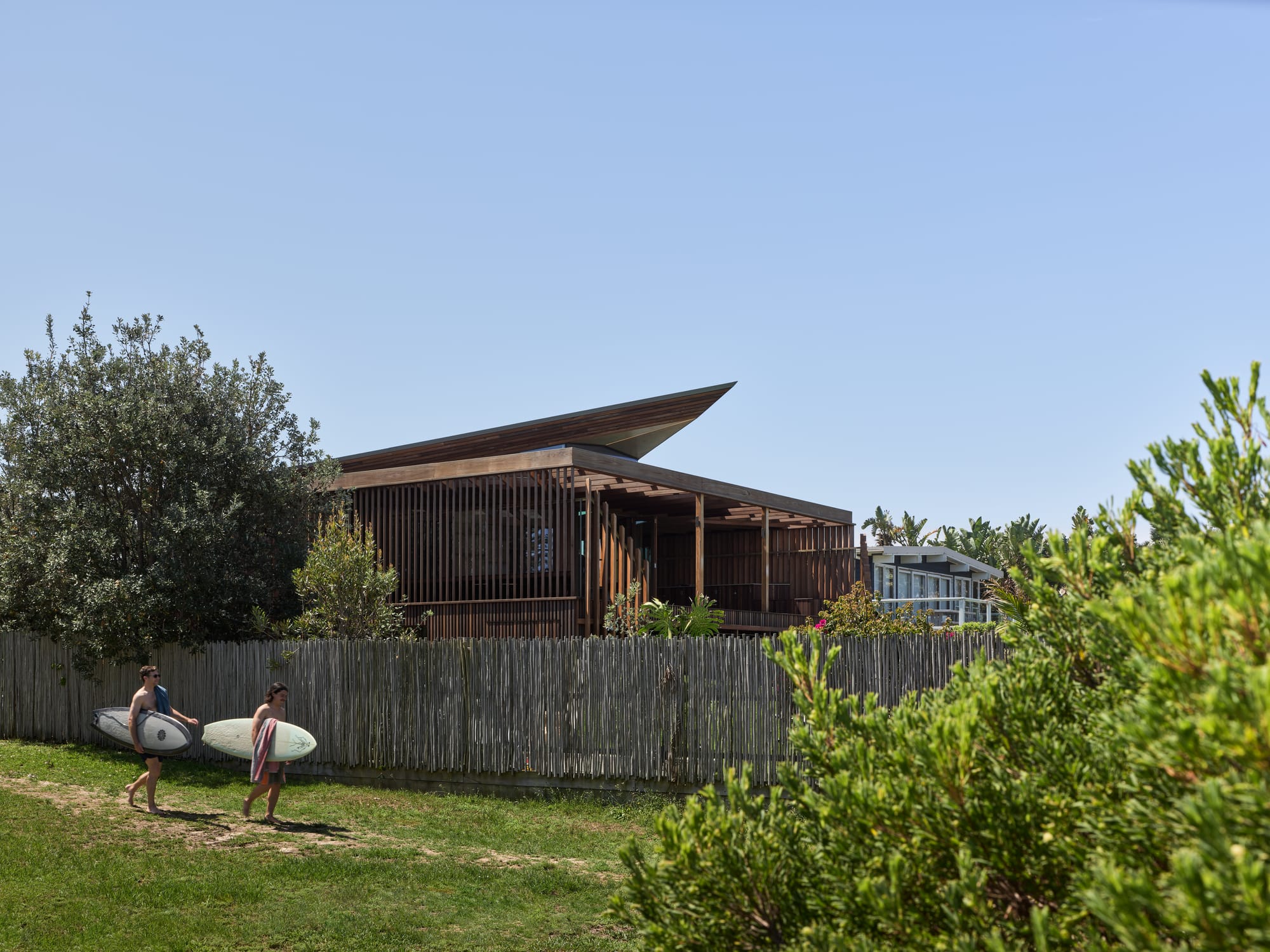 Cake House by Alexander Symes Architect. Photography by Barton Taylor. Two people with surfboards walk down a sandy path cutting across green grass, in front of a timber fence and timber clad home.