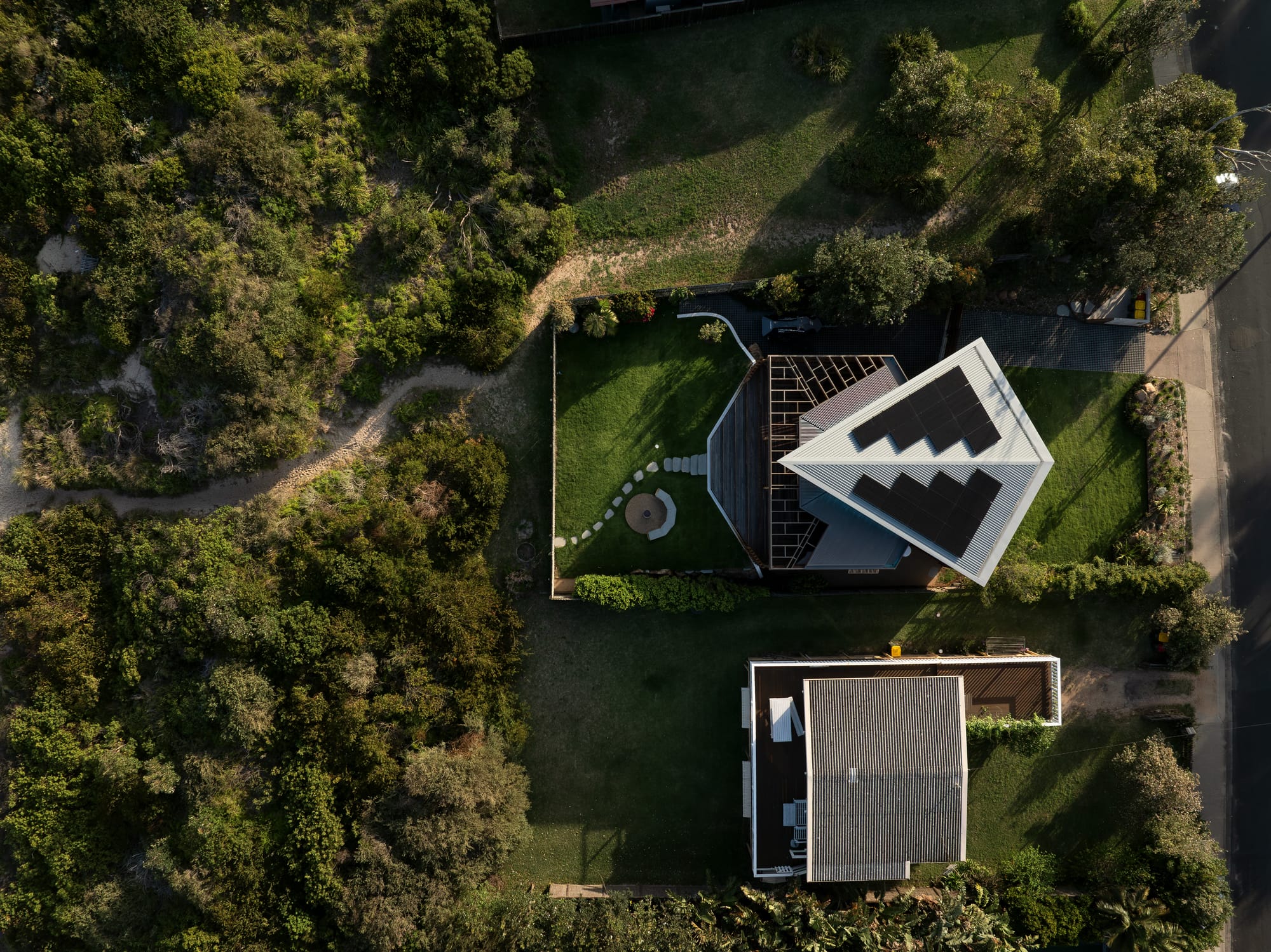 Cake House by Alexander Symes Architect. Photography by Barton Taylor. Aerial view of residential home with kite-shaped roof, grassy backyard and front yard and sand path leading through the scrub to the beach.