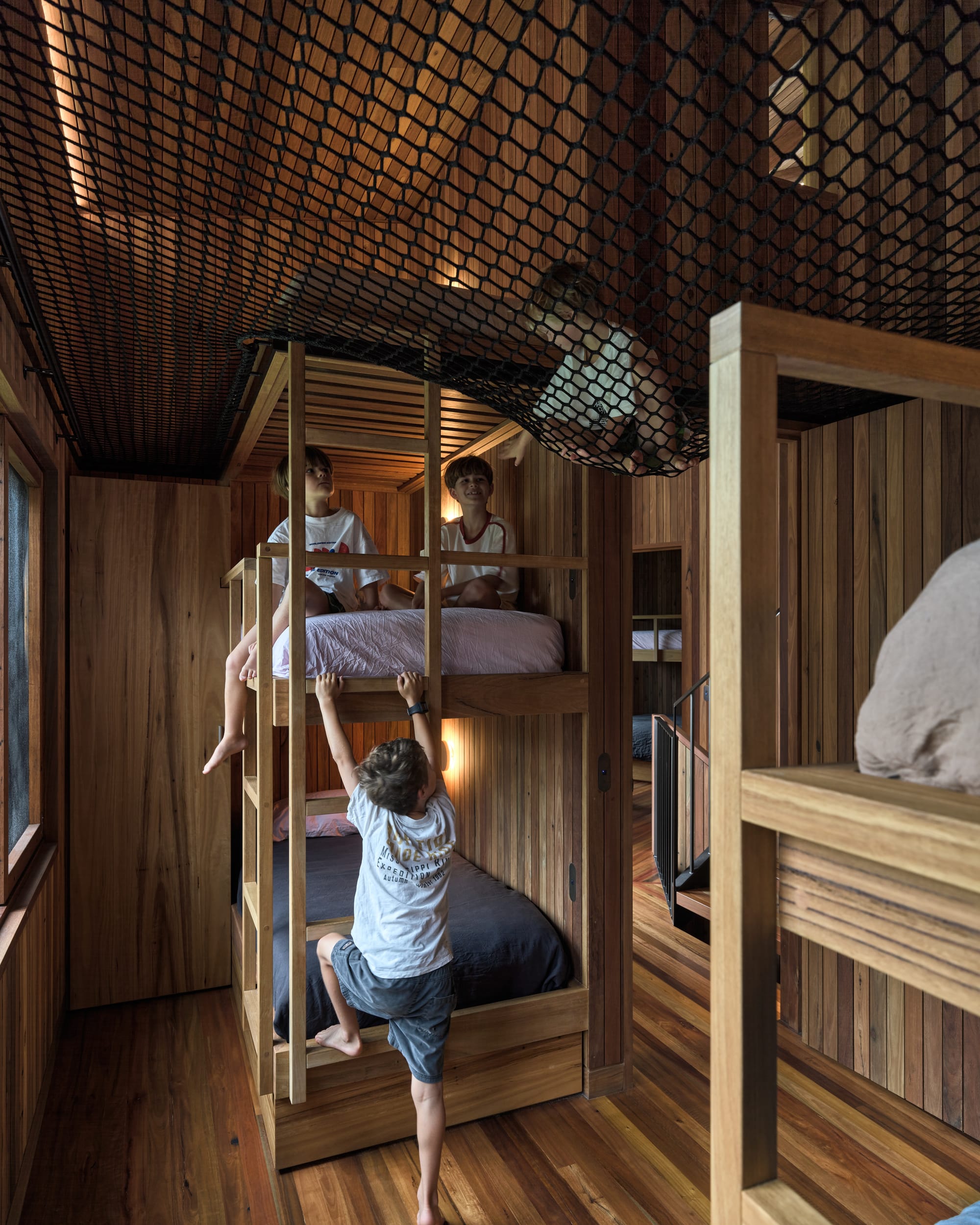 Cake House by Alexander Symes Architect. Photography by Barton Taylor. Childrens bedroom with timber floors, walls and bunk beds, and a netted ceiling, with multiple children playing.