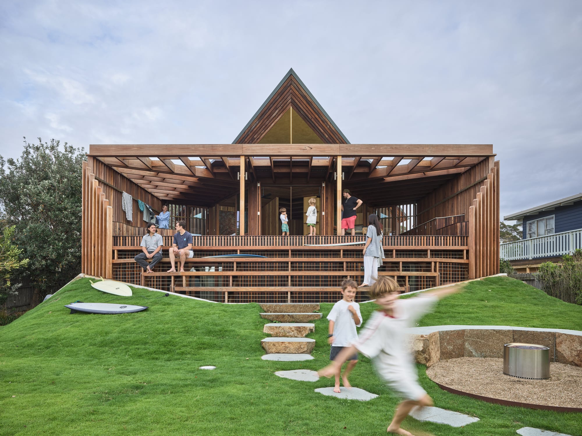 Cake House by Alexander Symes Architect. Photography by Barton Taylor. Rear facade of timber clad home with stairs and pitched roof, with lots of kids running around and green grass. 