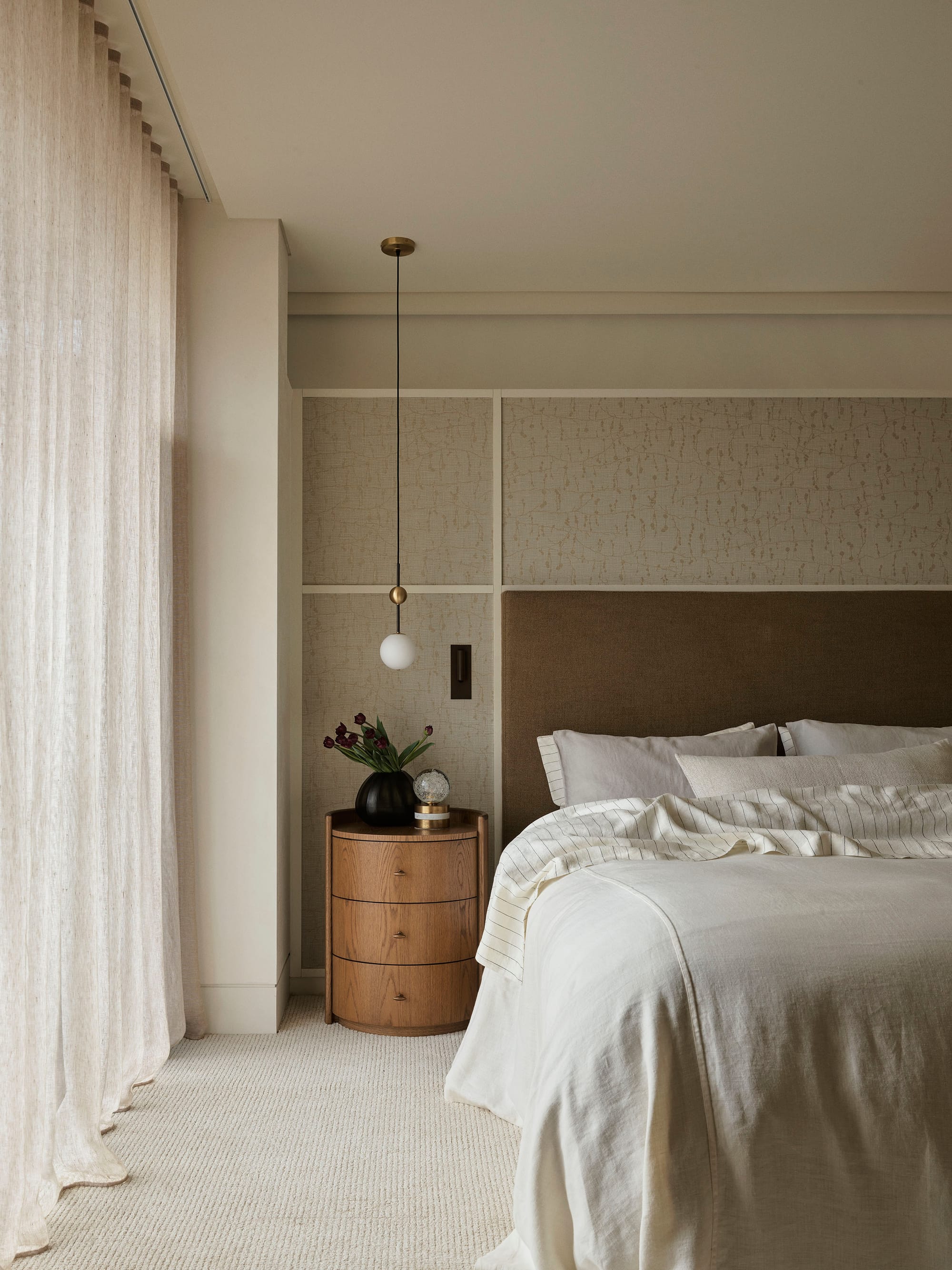 Bundarra Penthouse by Marylou Sobel Interior Design. Photography by Anson Smart. Bedroom with white bed linen, beige patterned wallpaper, full height white sheer curtains and circular timber side table.