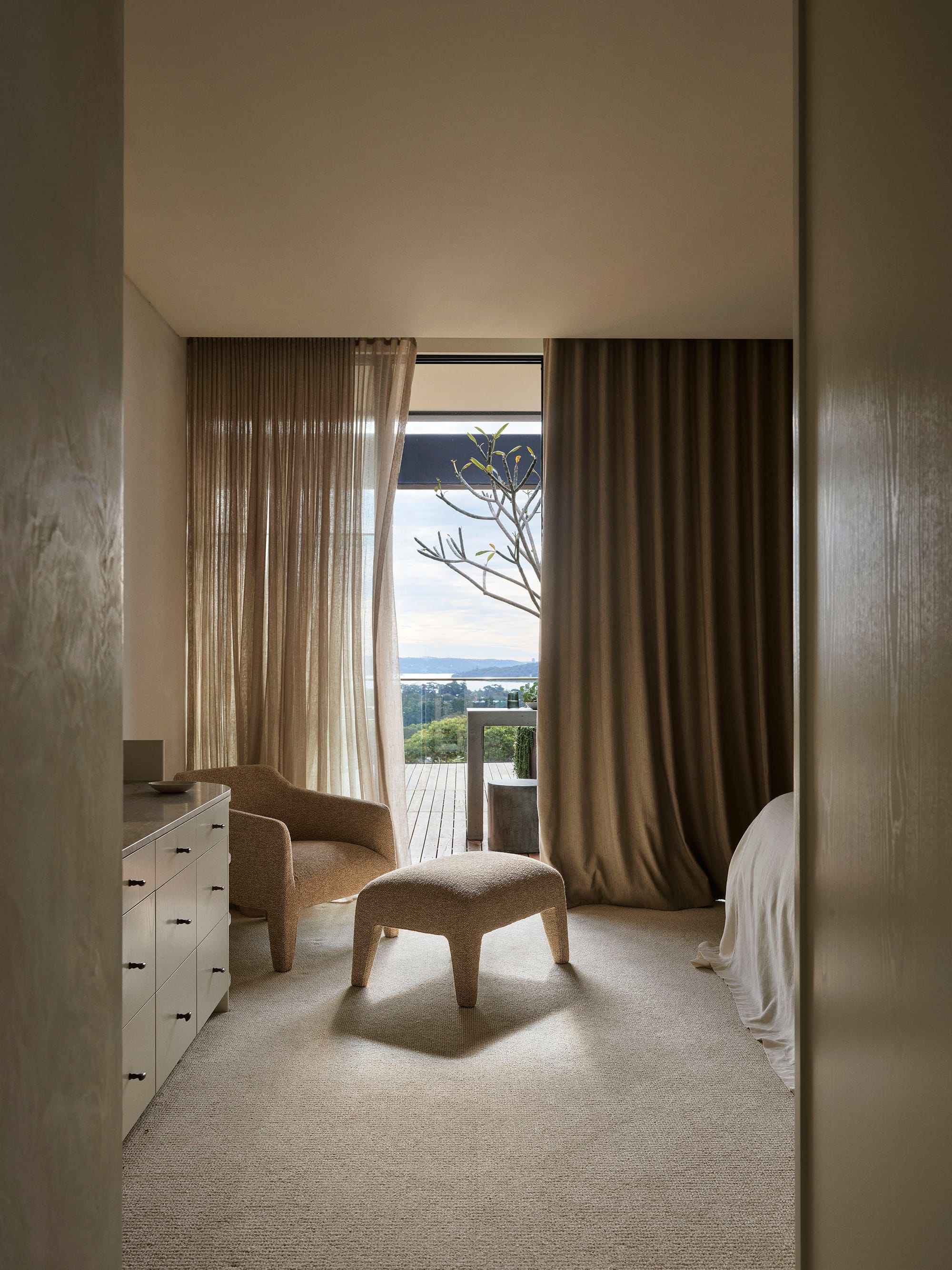 Bundarra Penthouse by Marylou Sobel Interior Design. Photography by Anson Smart. Bedroom with full height windows and doors opening onto deck with landscape views, cream carpets inside and a caramel armchair.