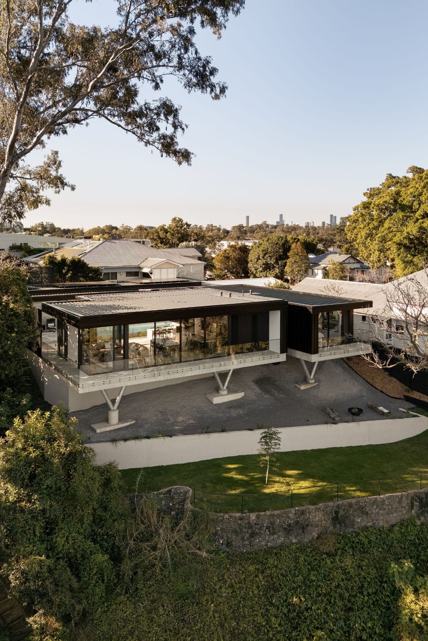 The River House, Indooroopilly by Sarah Waller Architecture. Photography by Alyne Media. Aerial image of residential home supported over sloped site, with flat black roof and balcony wrapping around the rear of the home.