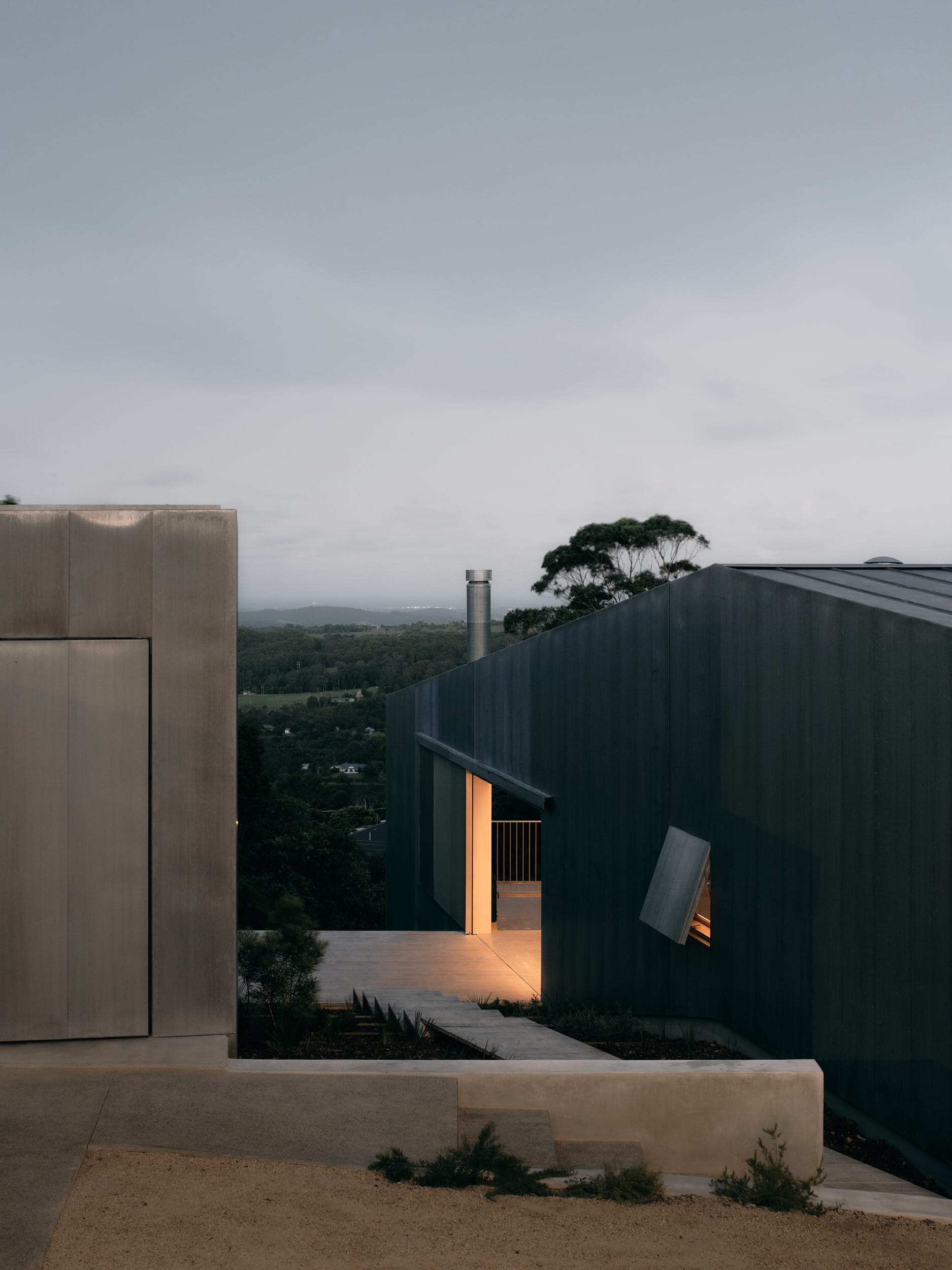 Mapleton House by Atelier Chen Hung. Photography by David Chatfield. Large zinc clad structure with opening, emitting war interior light, overlooking valley.