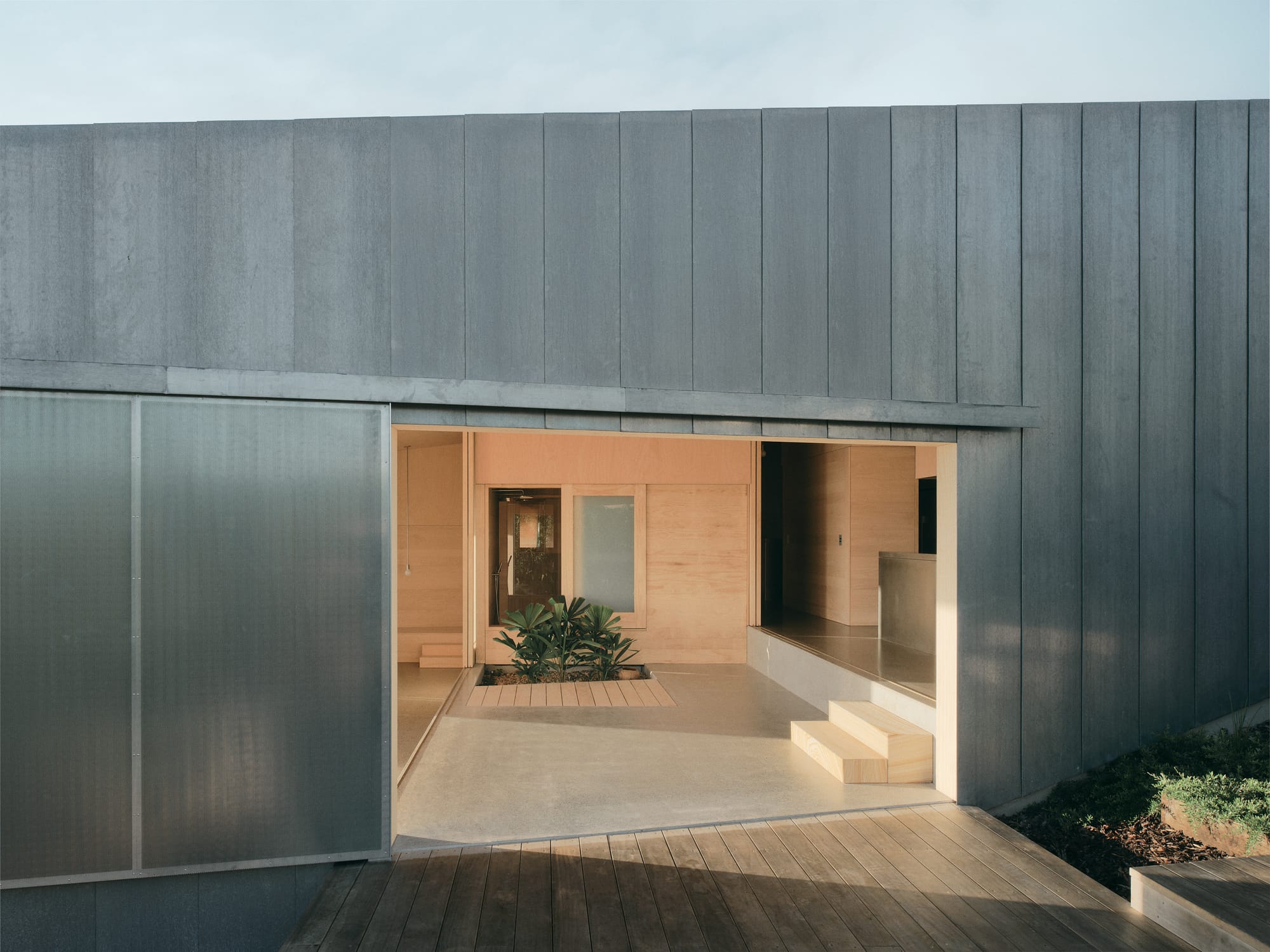 Mapleton House by Atelier Chen Hung. Photography by David Chatfield. Large opening in zinc-clad home, showing stepped interior with concrete floors and warm lighting.