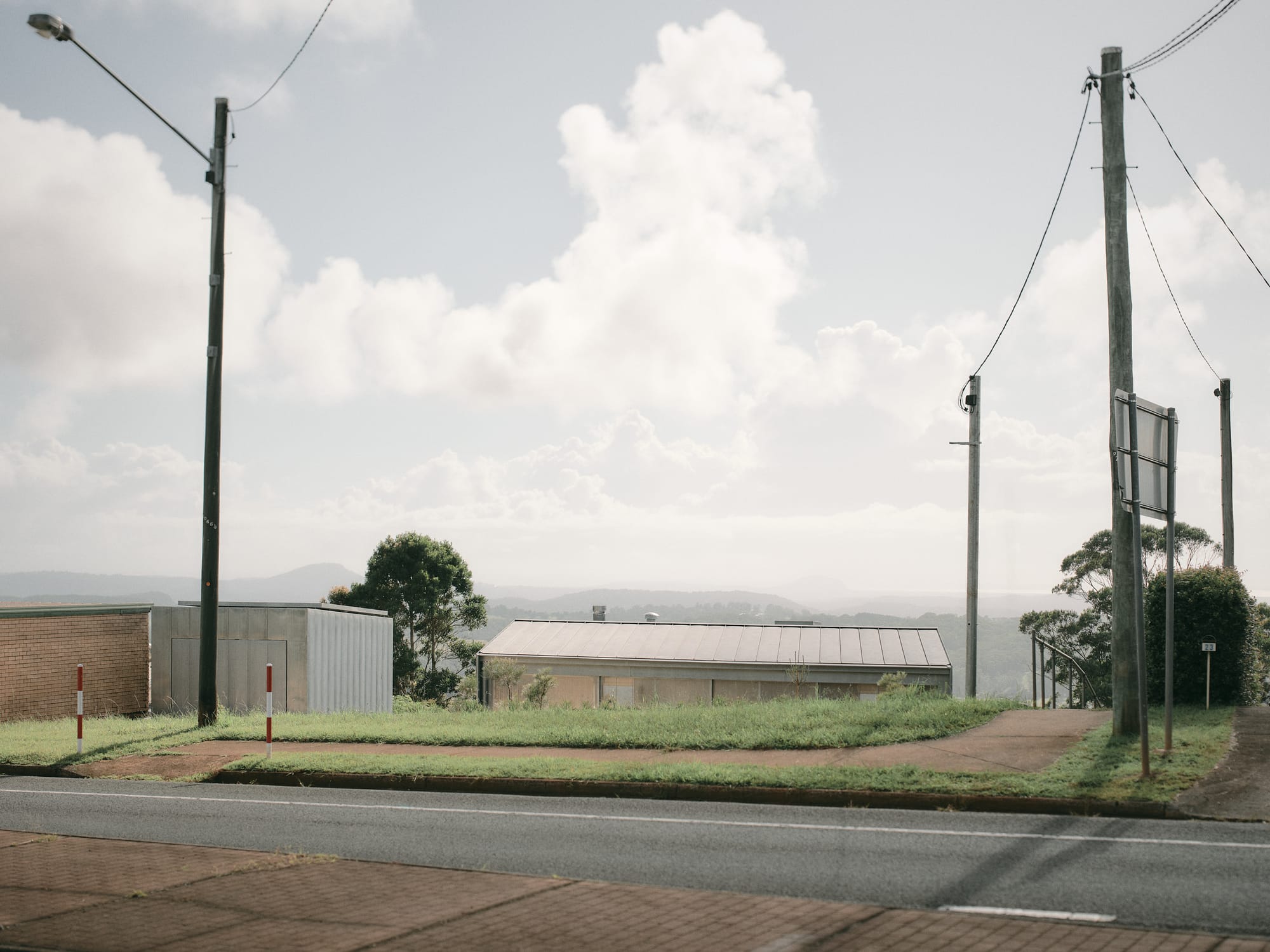 Mapleton House by Atelier Chen Hung. Photography by David Chatfield. Street with footpath cutting through grass and towards the horizon, with a roof in the midground and mountains in background.