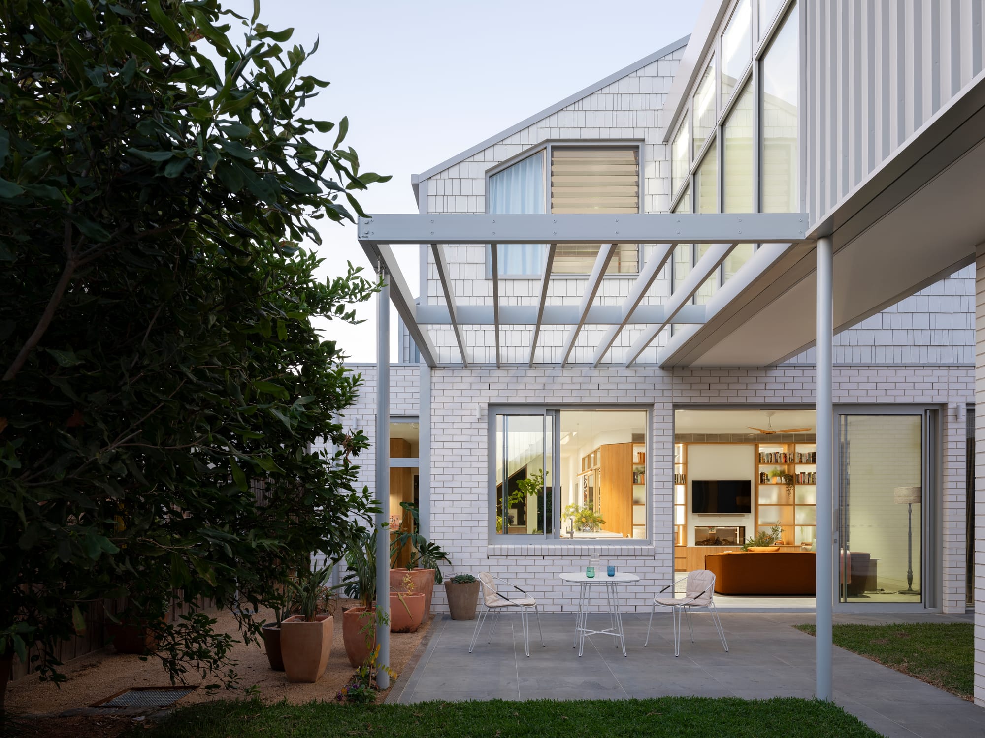Merrilands by BIJL Architecture. Photography by Tom Ferguson. Exterior facade with white brick and timber clad finish, exposed pergola courtyard with concrete flooring and large bush on left hand side.