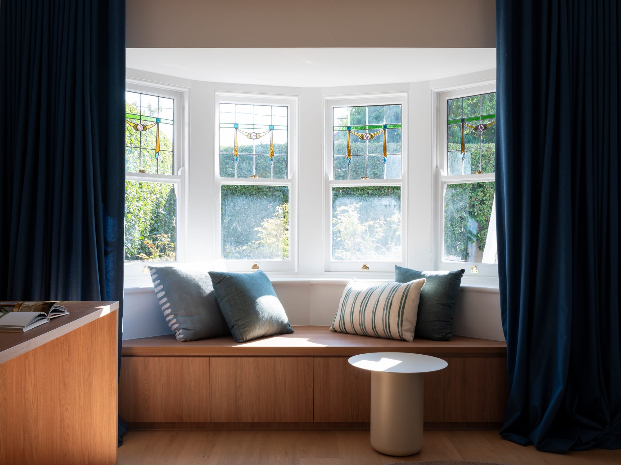 Merrilands by BIJL Architecture. Photography by Tom Ferguson. Bench window seat made from warm toned wood, with navy velvet curtains and white end table.