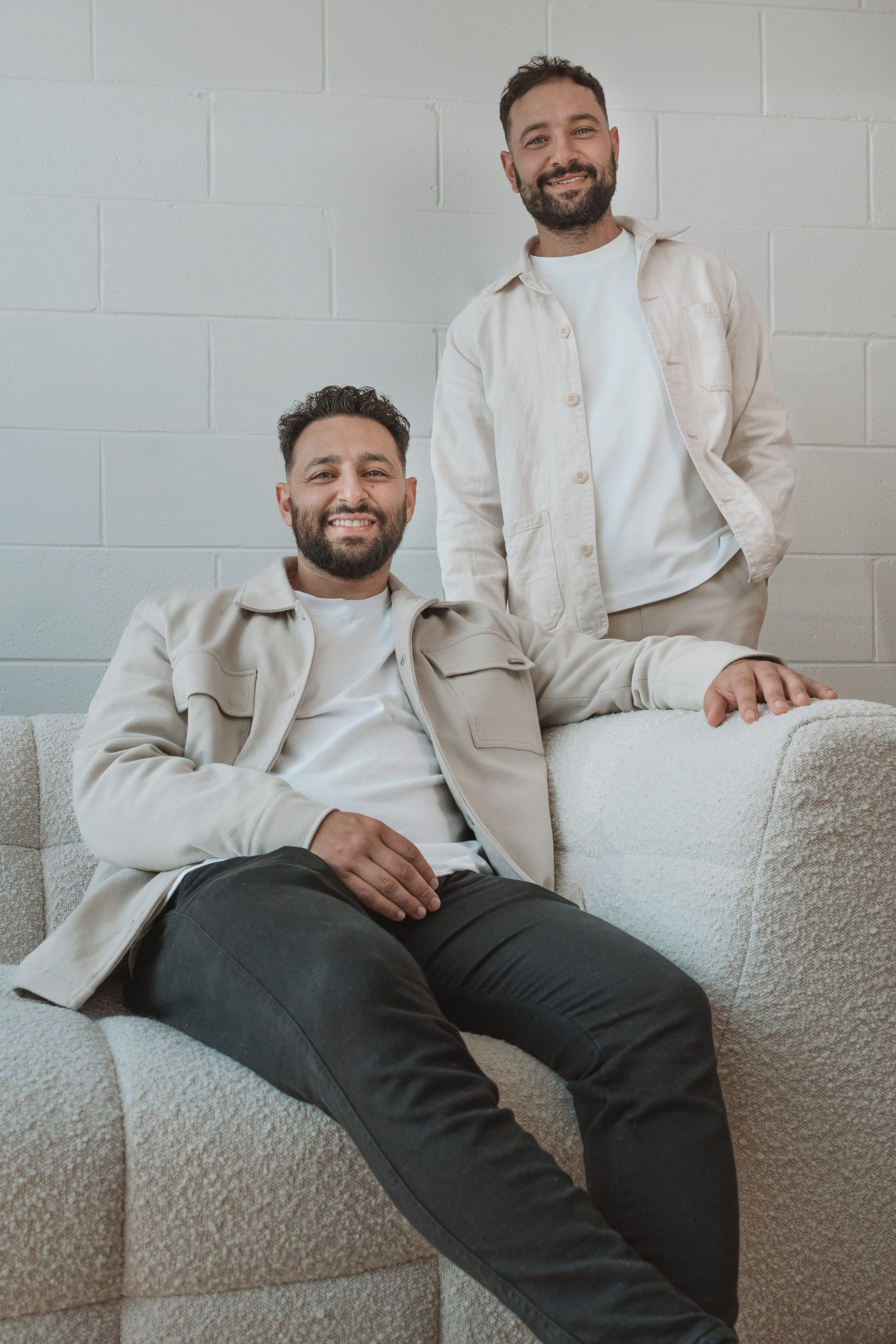 Dany and Mark Chalhoub of Prem.Built. Two men in beige outfits smiling, one sitting on couch and one standing behind. 