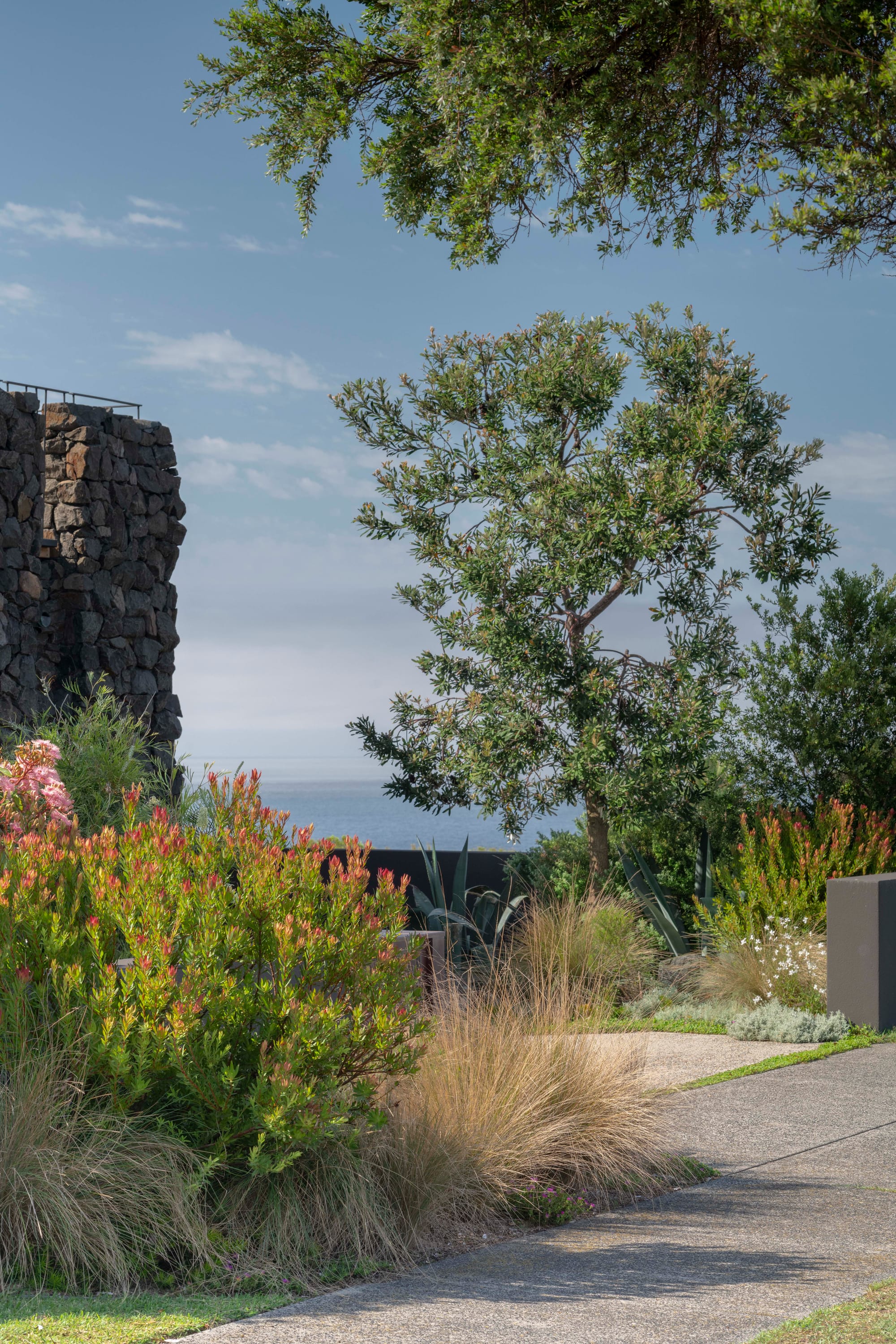 Stone House by Secret Gardens. Layered coastal planting with agave, native grasses and flowering shrubs beside a stone-clad wall.