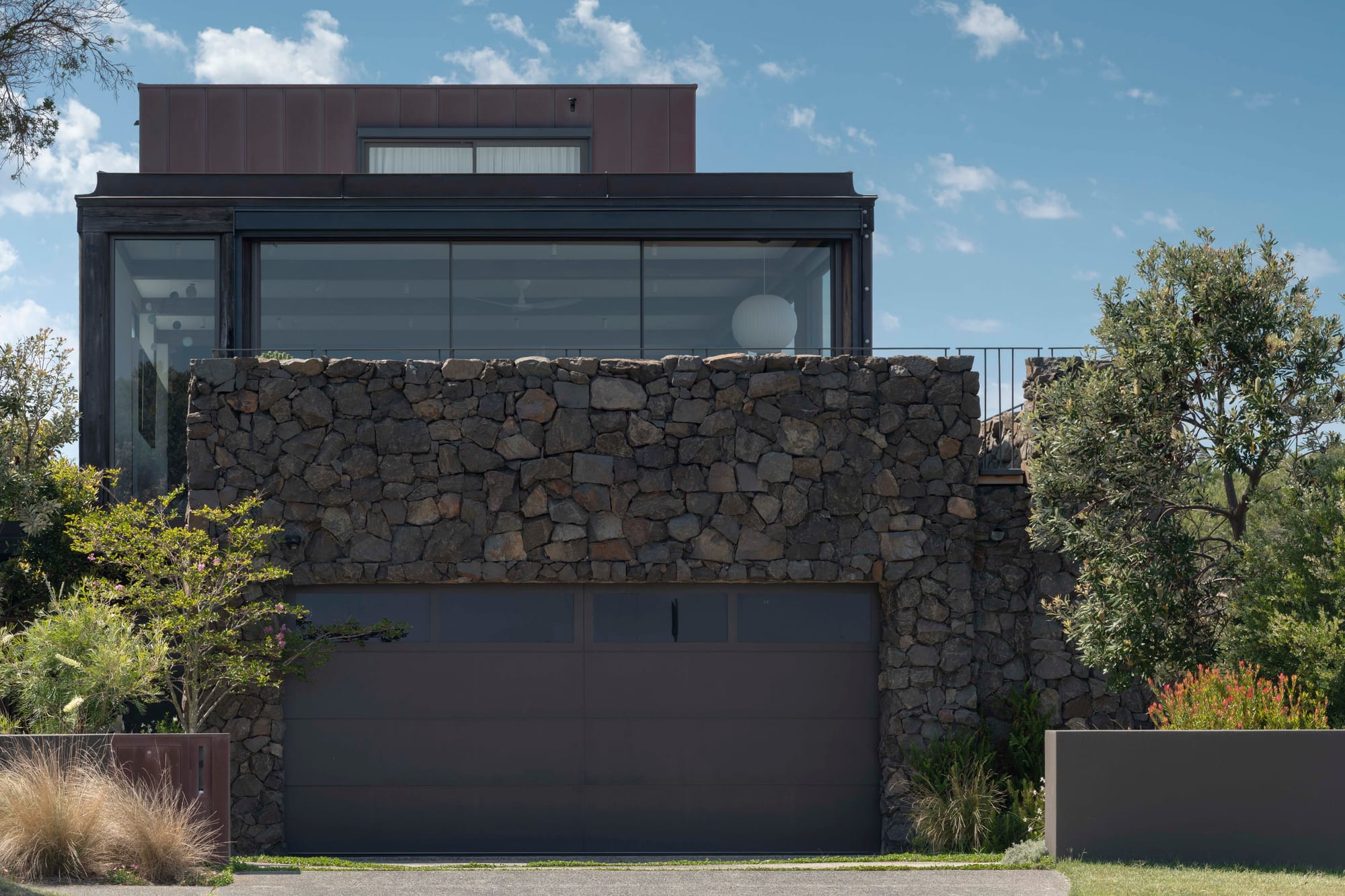 Stone House by Secret Gardens. Exterior view of Stone House framed by mature tea trees and coastal vegetation under a soft morning sky.