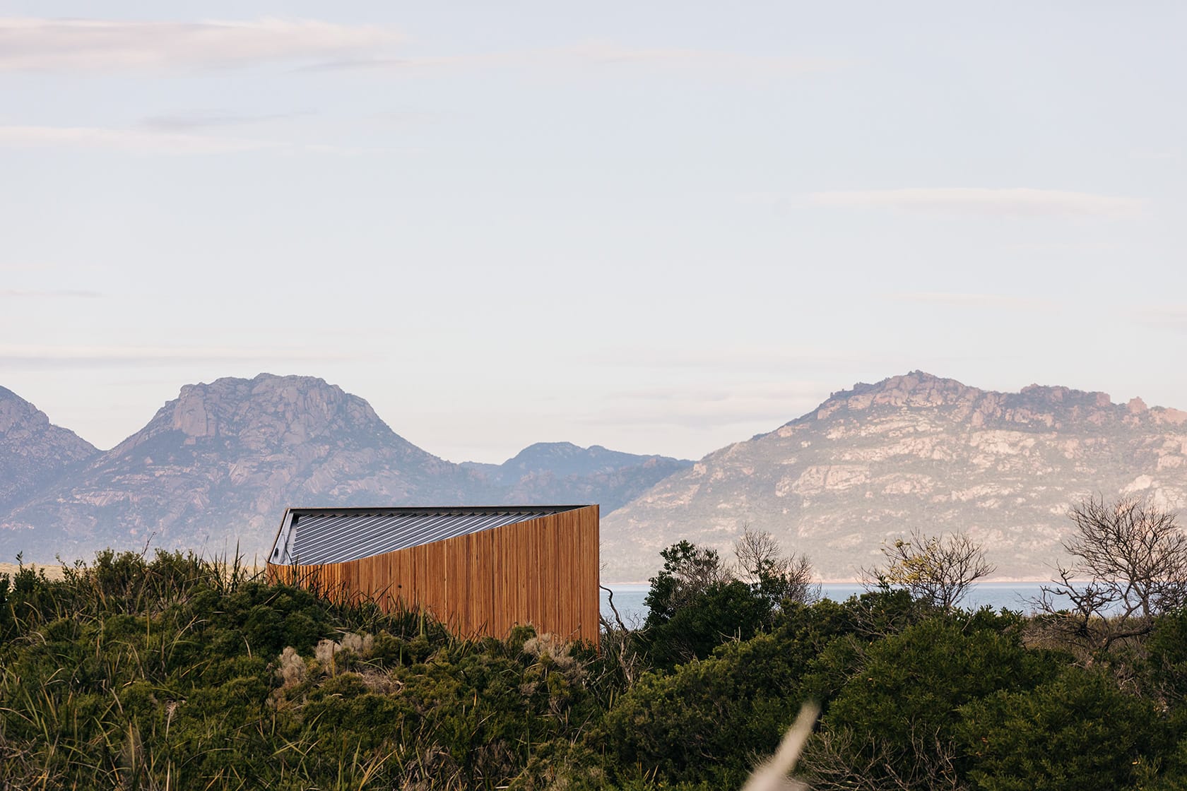 Studio Tasmania. Photography by Adam Gibson. Timber clad structure emerging from sand dunes and foliage, with ocean and hilly landscape in background.