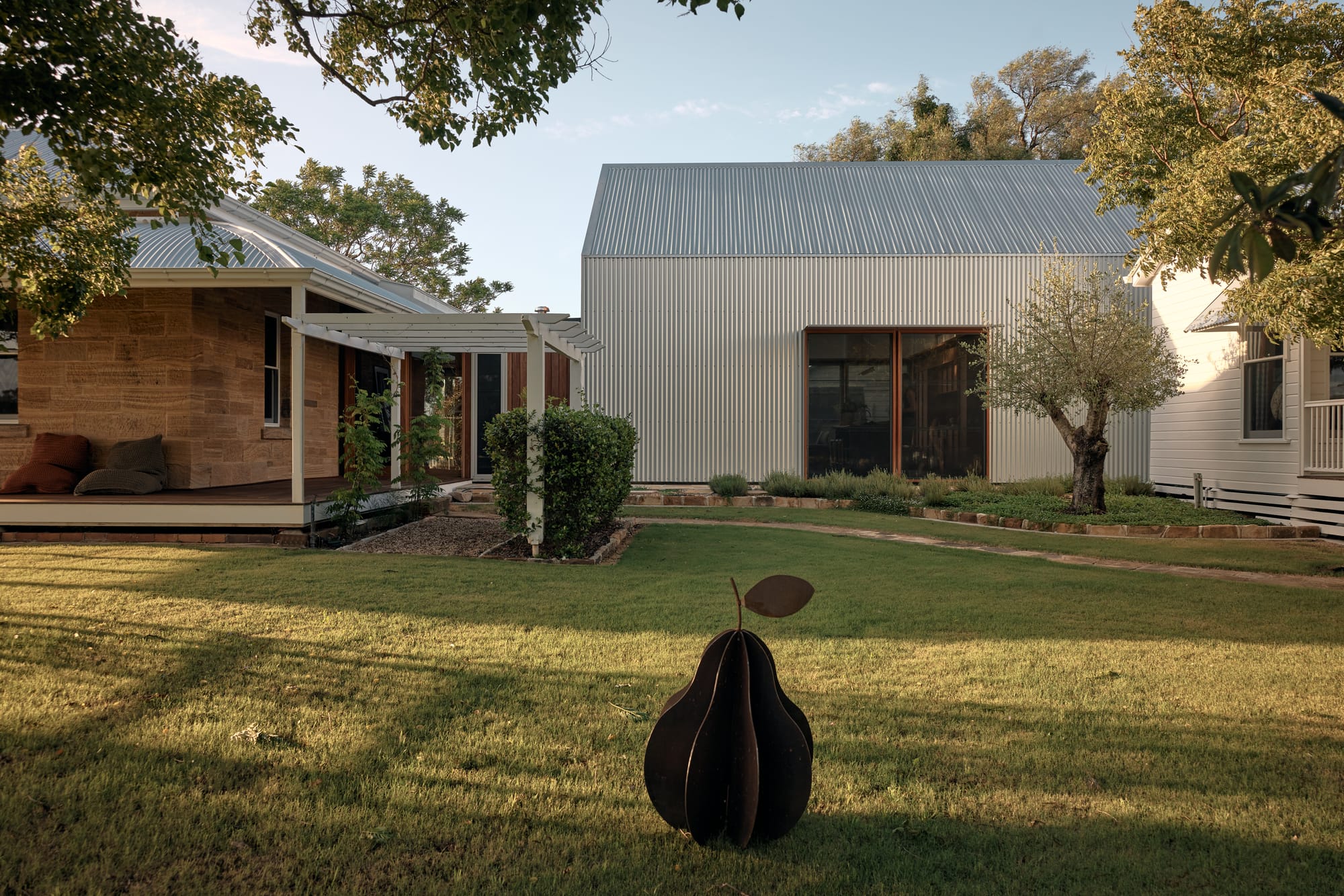 Binnum Homestead by Alexandra Buchanan Architecture. Photography by Cieran Murphy. Large grassed backyard with A-frame structure and traditional homestead in background, and metal pear sculpture in foreground.