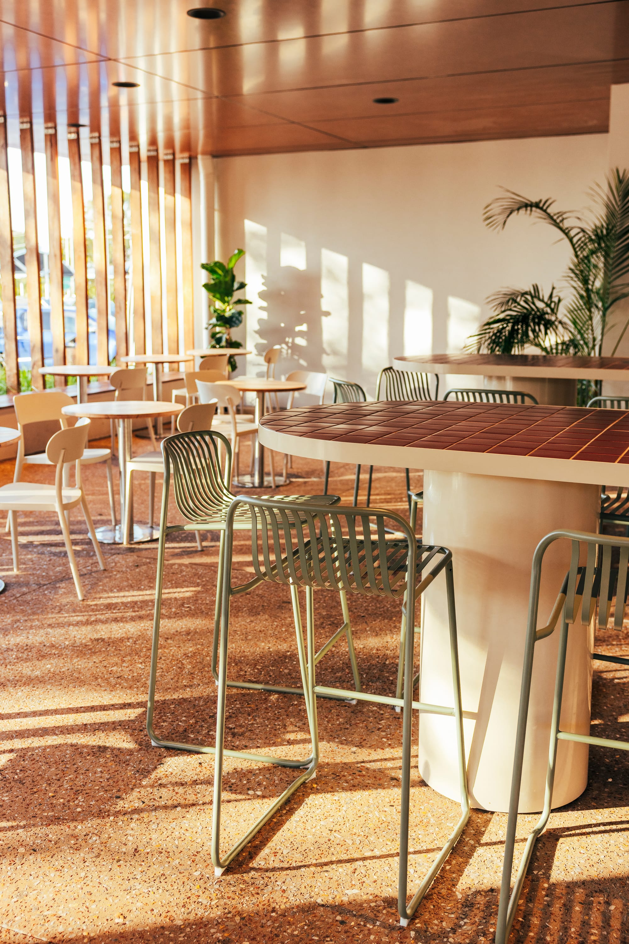 The Cafe by Bel.Bon Design. Photography by Sana Brotherson. Outdoor dining area with brown concrete floors, white steel bench seating with terracotta tiled countertops and green steel stools.