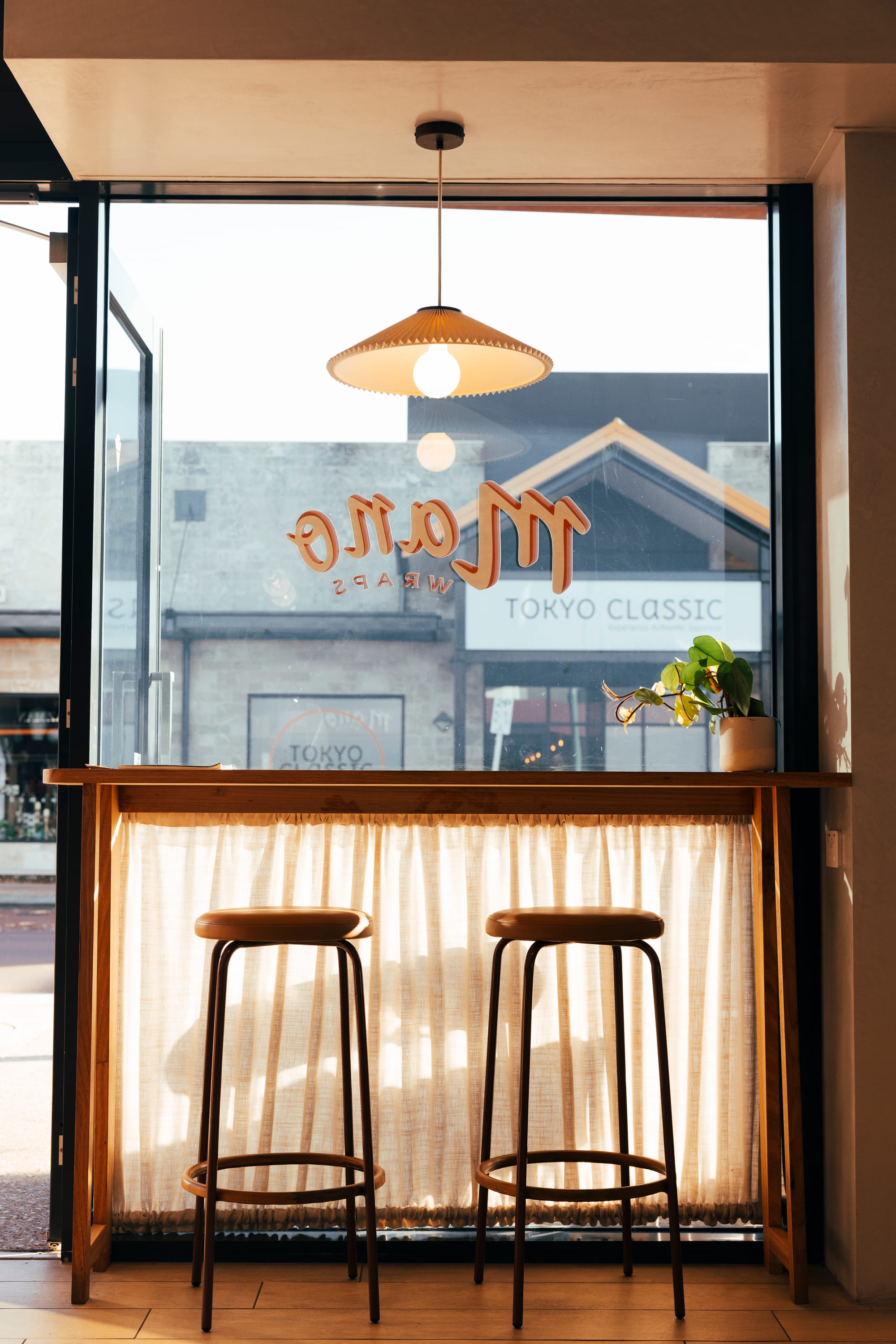 The Cafe by Bel.Bon Design. Photography by Sana Brotherson. Two stools in front of timber dench and full height window, with semi-sheer curtain.