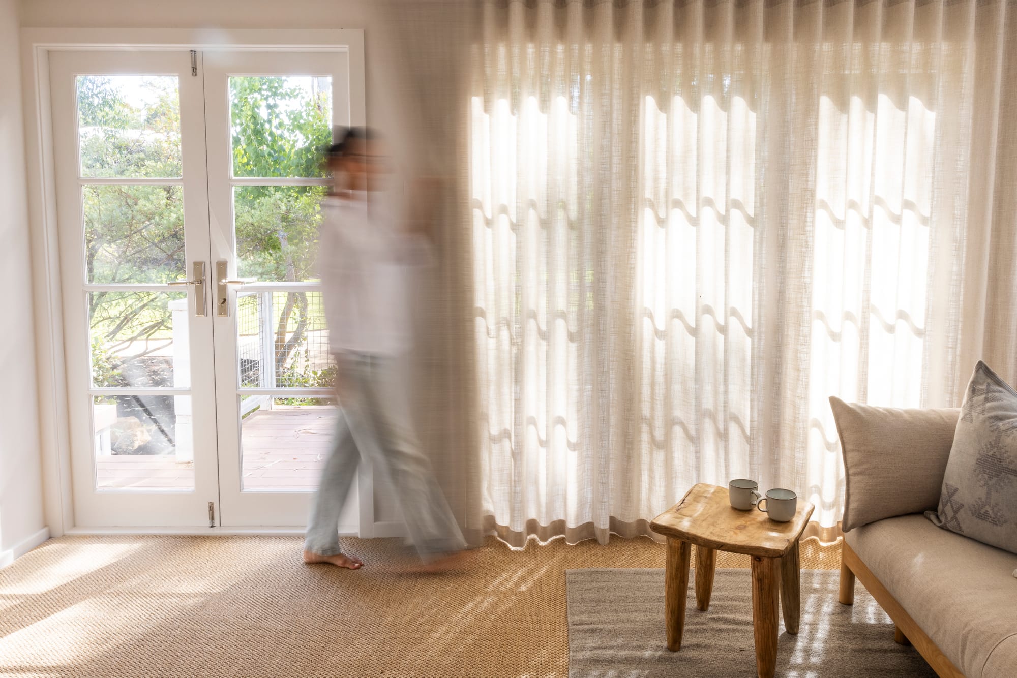The Cottage by Bel.Bon Design. Photography by Sana Brotherson. Living space with beige, jute carpet flooring, semi sheer linen curtains and white timber framed French doors.