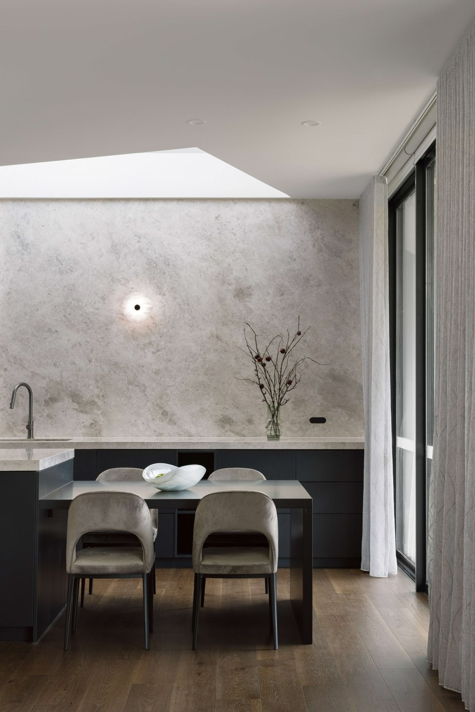 House on the Hill by ENCLAVE Architects. Photography by Elise Scott. Kitchen counter extending into shorter dining counter with dark cabinetry, grey upholstered velvet dining chairs and with dark timbe flooring and stone full height splashback.