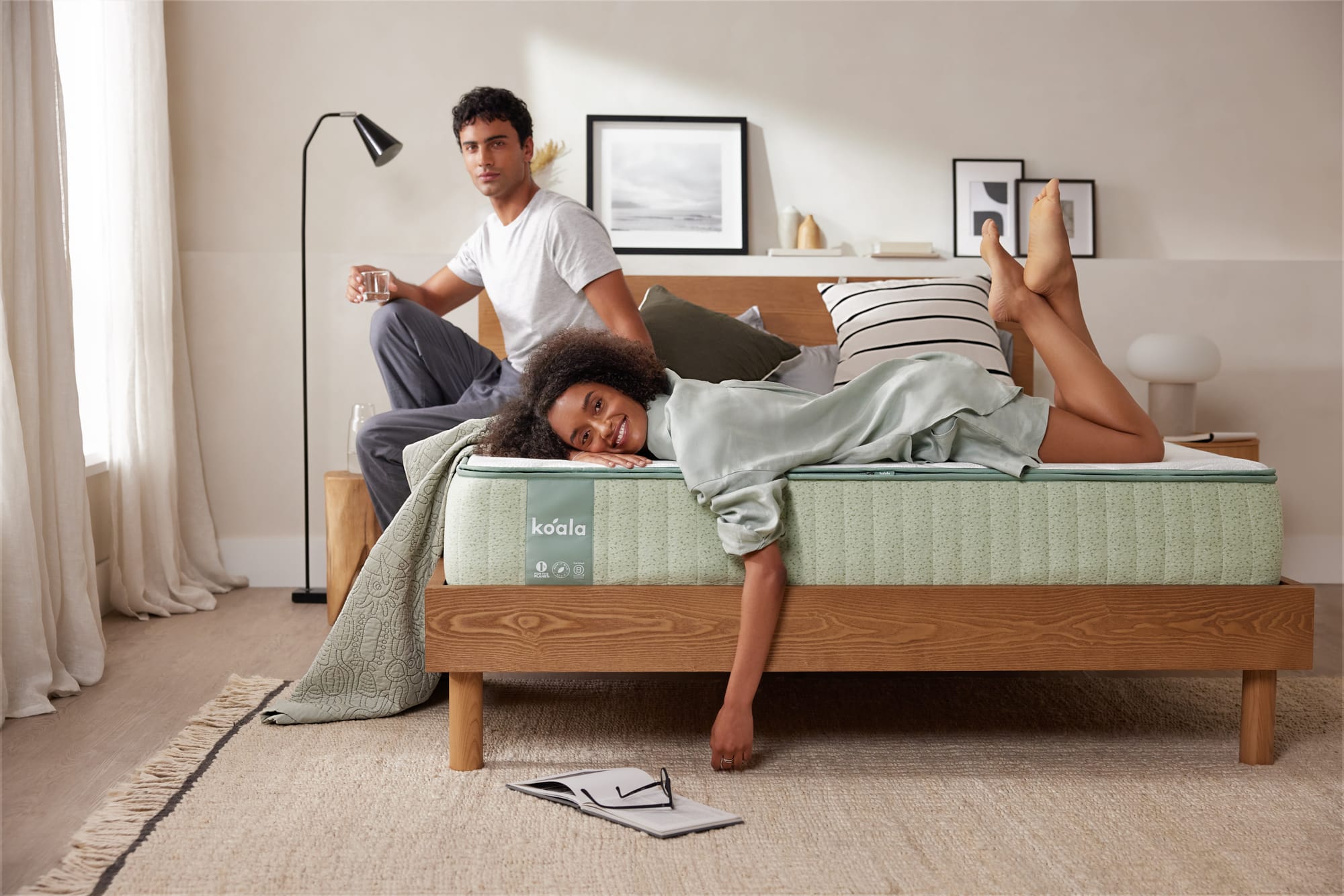 The Koala Mattress by Koala. Woman laying on bed smiling, with man sitting behind her, in room with jute floor rug and white walls.