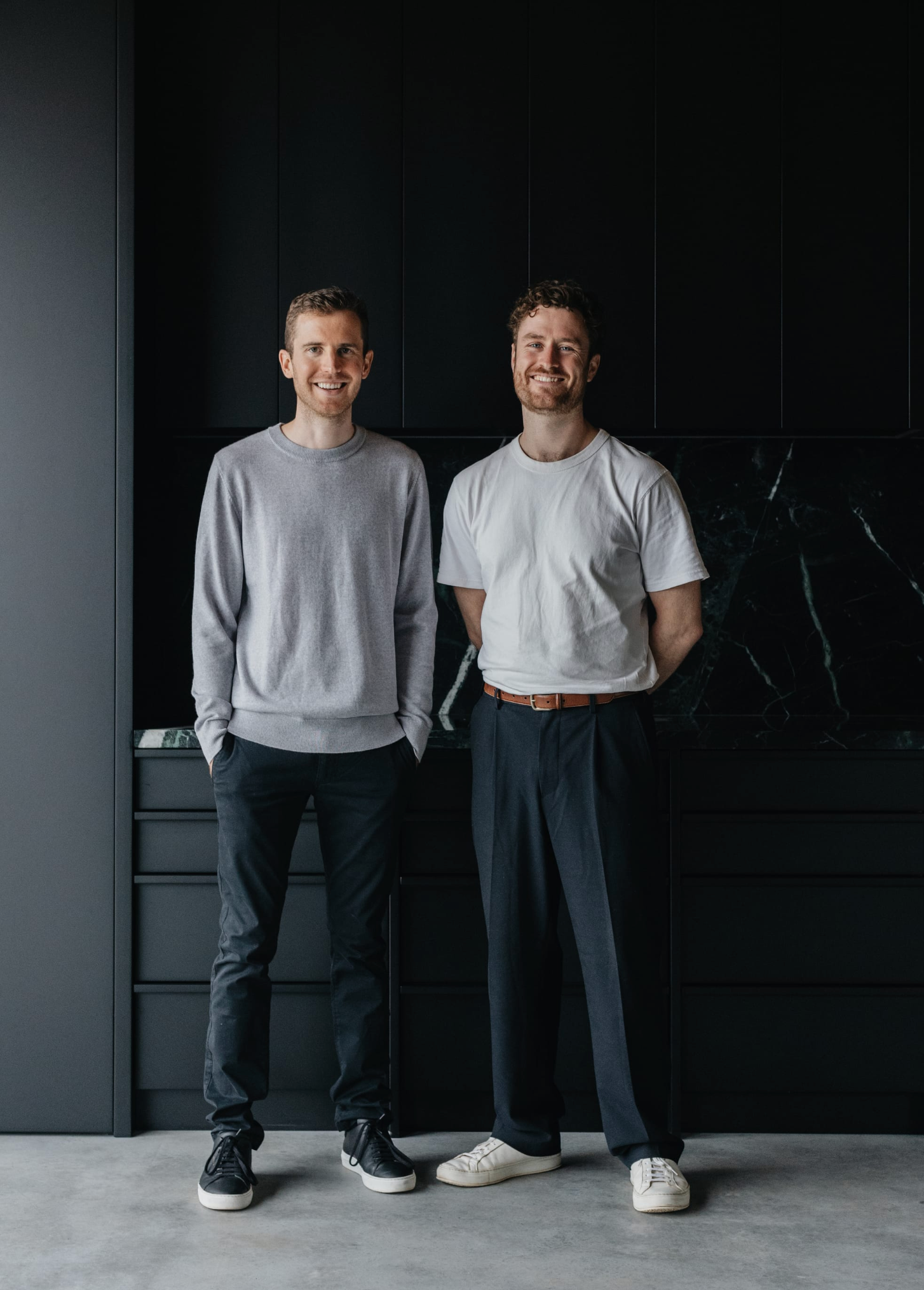 Liam Corr and Jeffrey Baikie of Baikie Corr Architecture. Two men stand in front of black wall in dark, neutral outfits. 