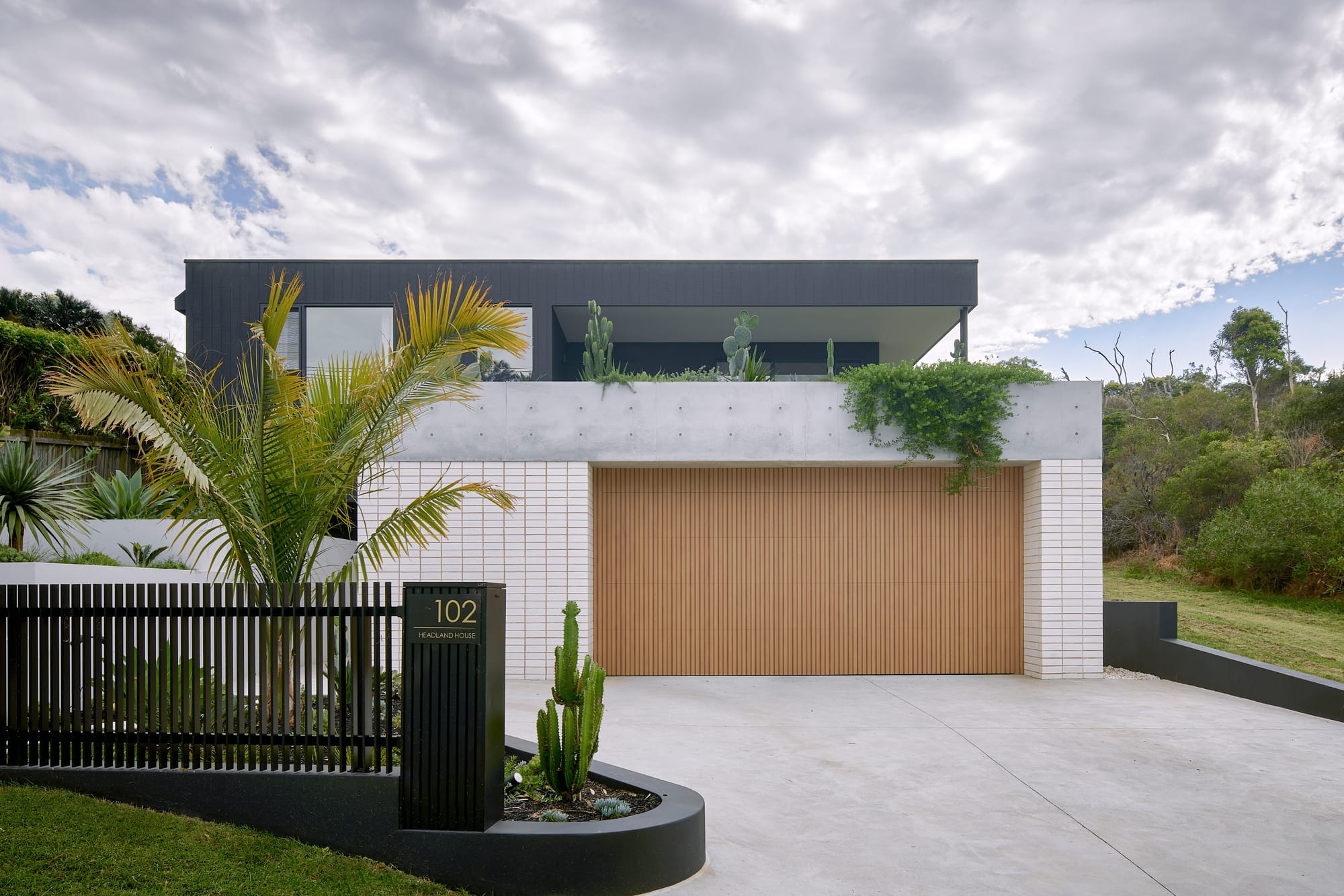 Headland House by Baikie Corr Architecture. Photography by Toby Peet. Front facade of home with timber garage door, concrete driveway, white brick ground floor, black upper level and large concrete balcony with hanging plants. 