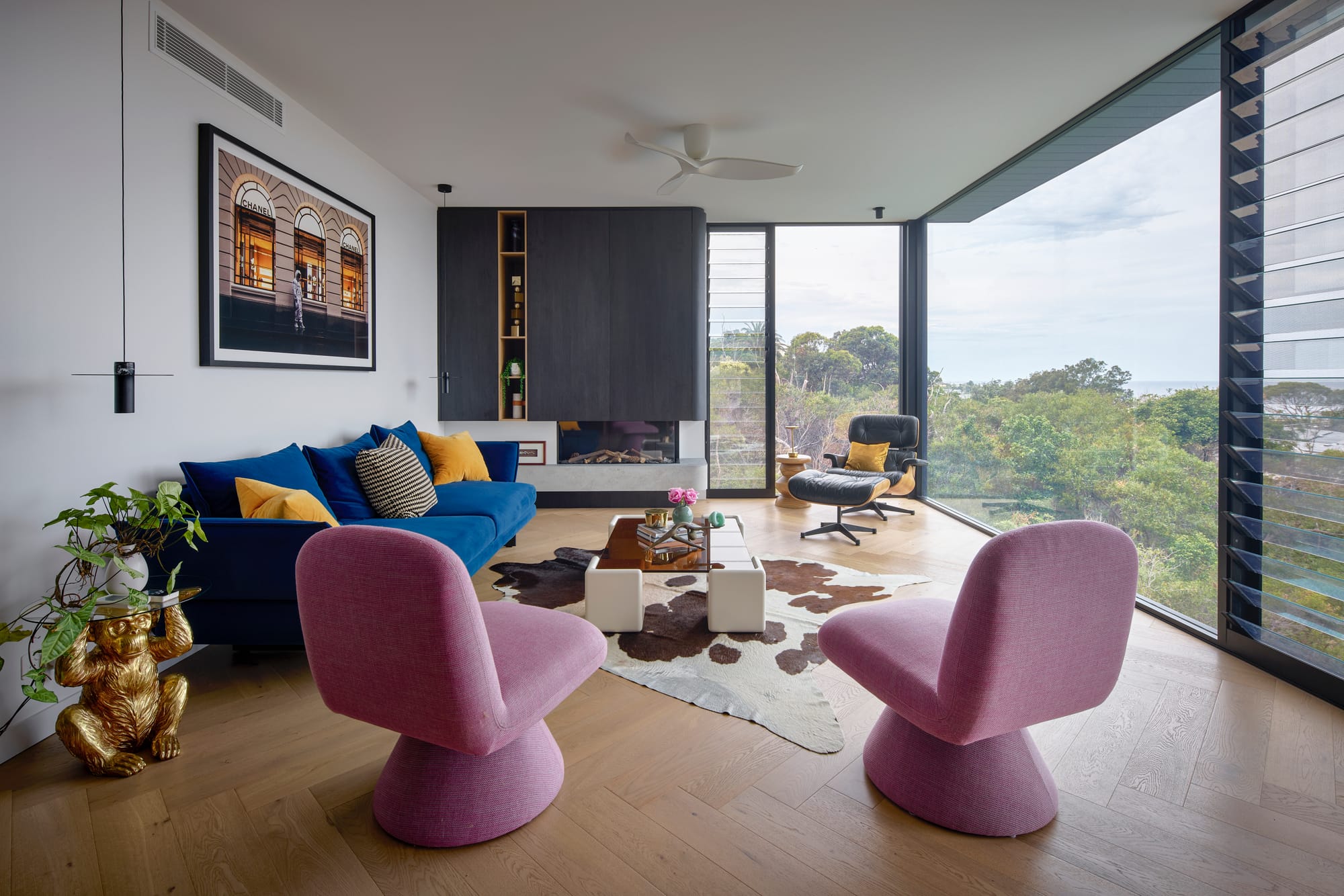 Headland House by Baikie Corr Architecture. Photography by Toby Peet. Eclectic living room with full height glass windows, herringbone timber floors, pink armchairs, a blue couch, cowhide rug and black fireplace. 