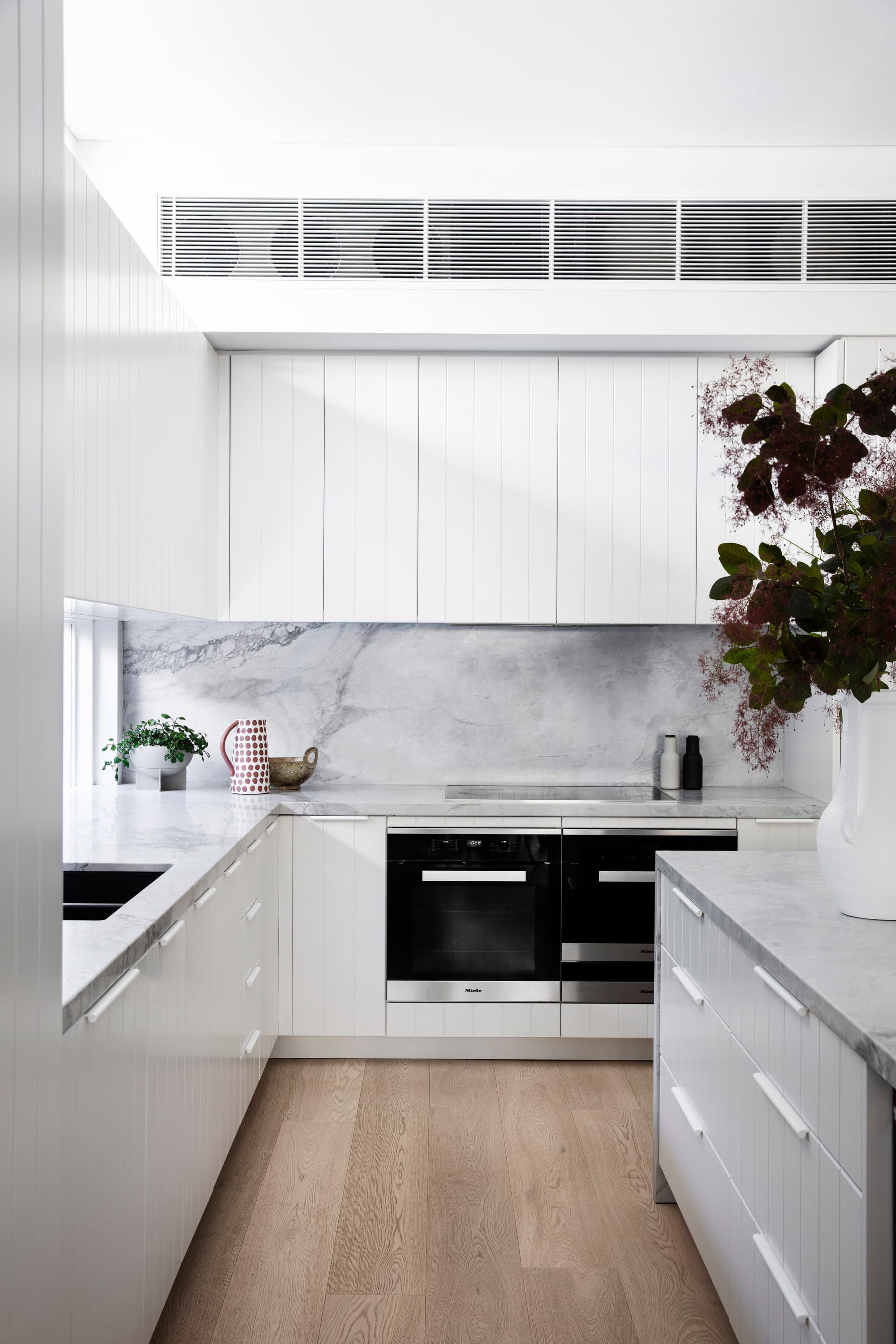 Bondi Beach House by Baikie Corr Architecture. Photography by Chris Warnes. White kitchen with timber floors, stone countertop and splashback, large floral arrangement in white vase on countertop and black appliances. 