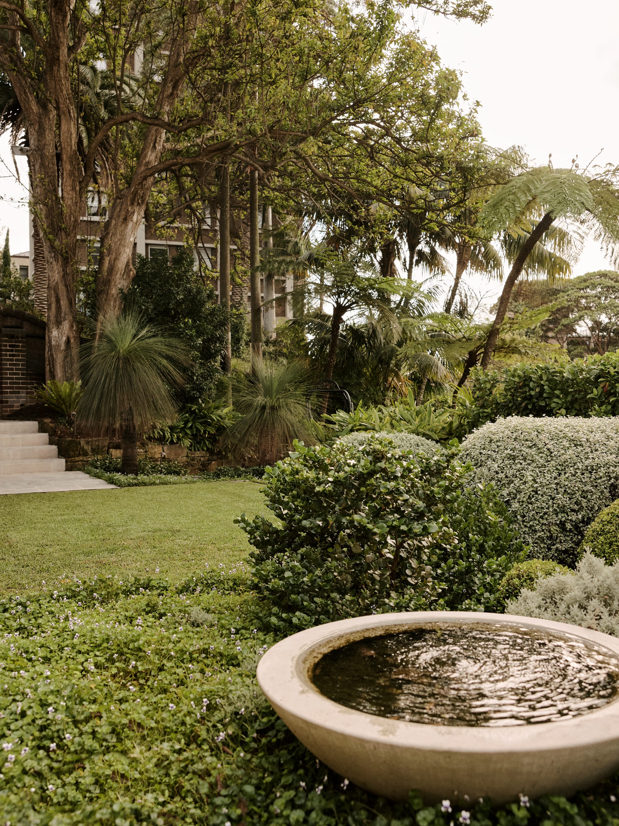 The Darling Edit by Marylou Sobel Interior Design. Photography by Anson Smart. Garden with round concrete water fountain on floor, low shrubs and ground cover, and stairs in background.