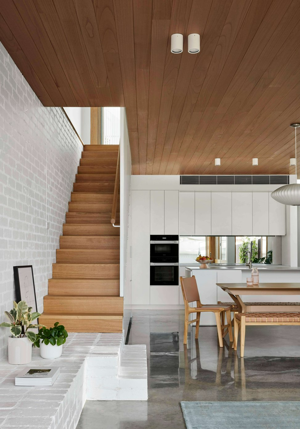 Rosie by Eckersley Architects. Photography by Tess Kelly. Open plan kitchen and dining space with polished concrete floors, white brick walls, a timber staircase and white cabinetry. 